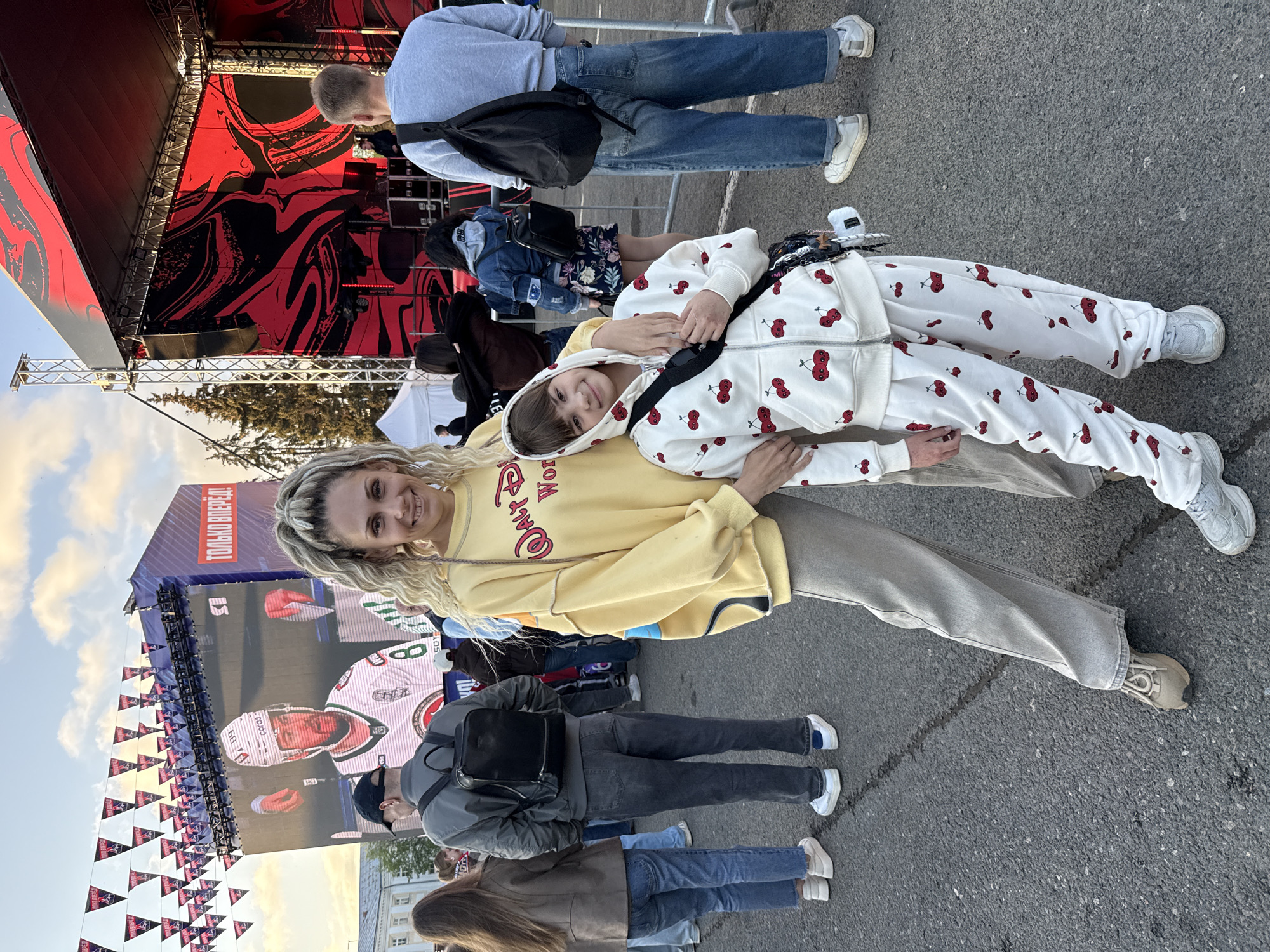 The image shows a mother and child posing for a picture at an outdoor event in Заволжье, Russia. The mother, with blonde dreadlocks, wears a yellow sweatshirt and gray pants. The child, wearing a white cherry-patterned tracksuit, is lying on the mother, embracing her. Behind them is a crowd of people, a large screen showing a hockey player, and red pennants decorating the area. A stage with a red and black backdrop is visible in the background. The ground is asphalt.