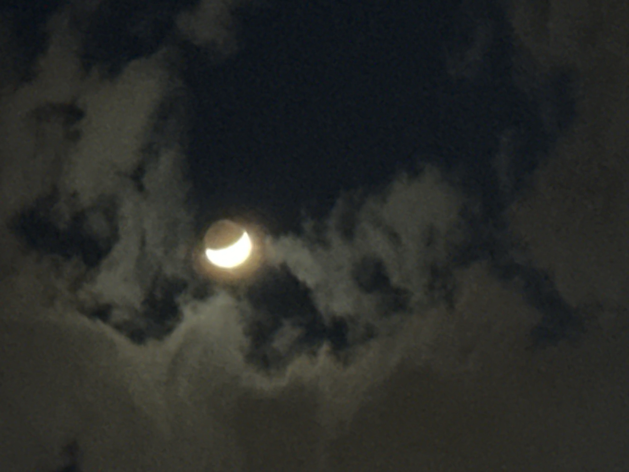 A partial lunar eclipse is visible in a dark, cloudy sky. The moon appears as a crescent shape, illuminated on the right side. The clouds are dark and patchy, obscuring parts of the sky. The scene is a nighttime sky, likely outdoors, with no visible buildings or landmarks that would indicate a specific location within Tallinn, Estonia. There are no people or specific objects in the image. The overall impression is of a celestial event observed through atmospheric conditions.