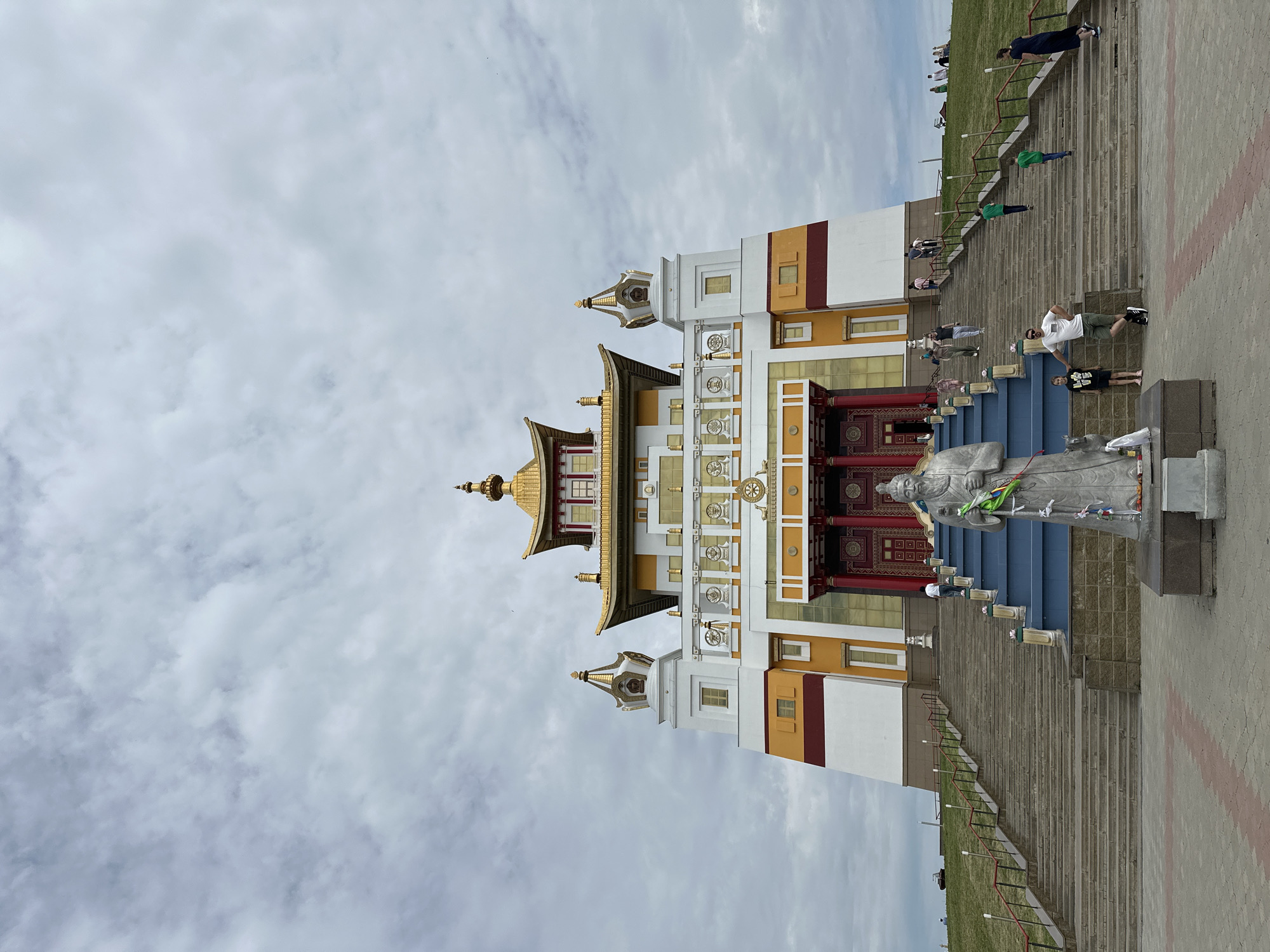 In Elista, Russia, a wide stone staircase leads up to the golden-roofed Buddhist temple known as the Golden Abode of the Buddha Shakyamuni. A gray stone statue of a robed figure holding a staff stands at the foot of the stairs. Several people are visible on the stairs and the grassy hill to the right, some walking up the steps, others standing and observing. The sky above is cloudy.