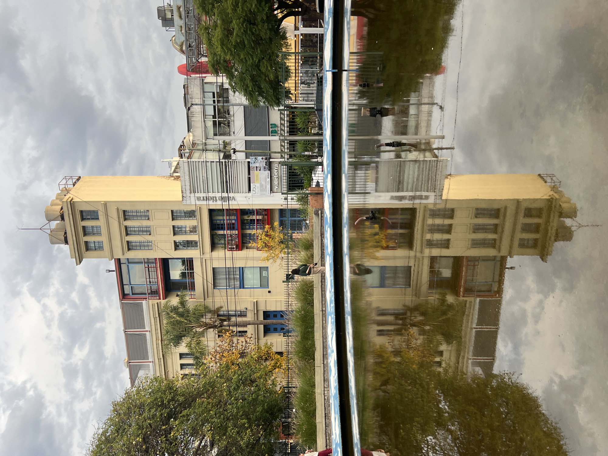 The image shows a reflection of buildings, trees, and sky in a body of water, likely a fountain or pool. The buildings are a mix of architectural styles, with one prominent yellow building having multiple stories and an elevated structure on top. There's a mix of residential and commercial buildings, including a building with "valle tierra" sign. The sky is cloudy and overcast. In the foreground, there is a metal rail of the pool. A person standing by the pool is also visible in the image. The location is identifiable as the area around Plaza Mafalda in Buenos Aires, Argentina.