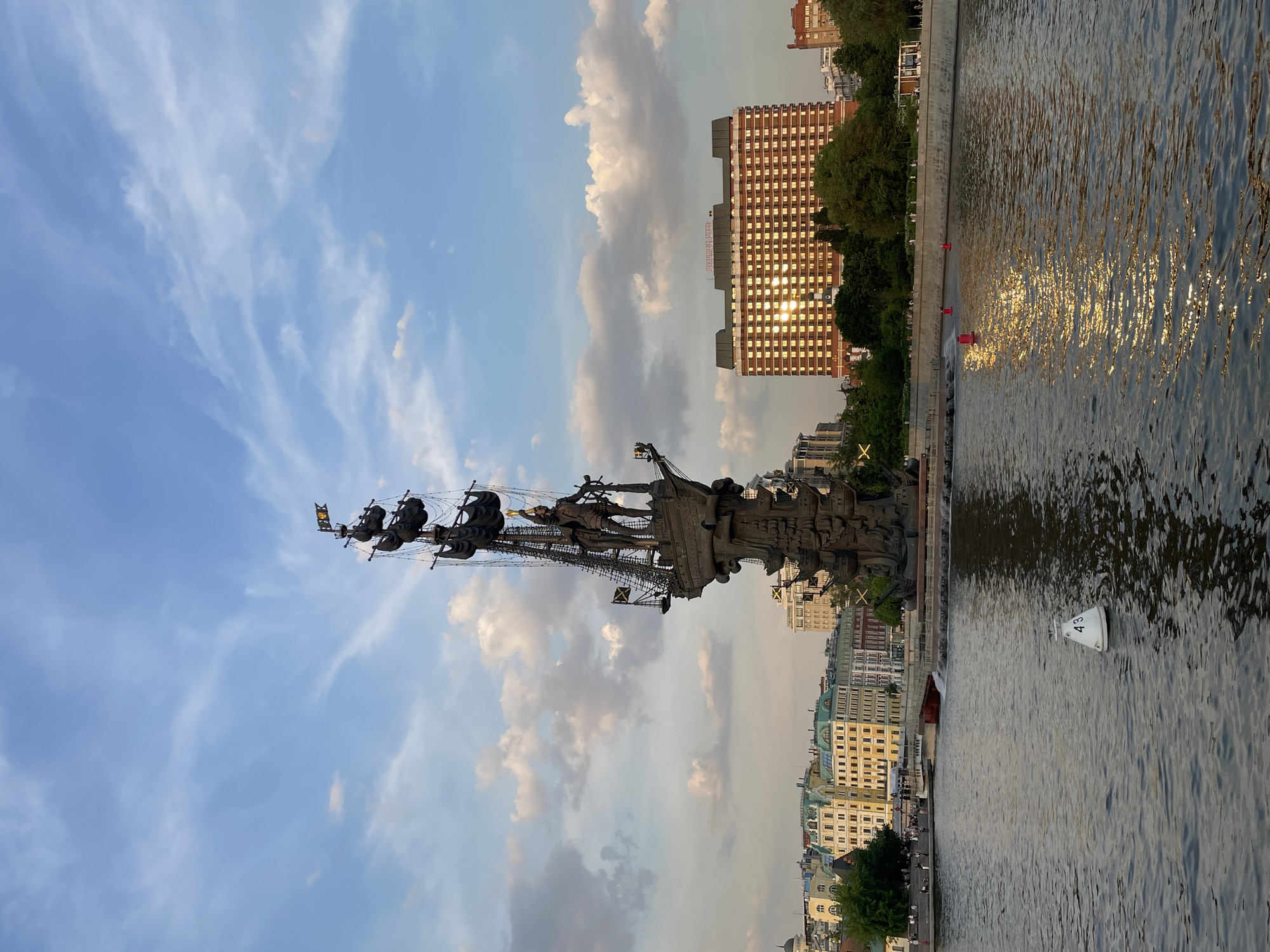 This image captures a panoramic view of the Peter the Great Monument in Moscow, Russia, situated on the Moskva River. The monument, crafted from bronze, depicts Peter the Great standing on a tall sailing ship, holding a helm. The intricate structure of the ship and its base are visible, featuring multiple figures and elements.
The scene is set during the late afternoon or early evening, indicated by the warm, golden light of the sun reflecting off the water and buildings. The sky above is a clear blue with scattered, wispy white clouds, suggesting pleasant weather.
The Moskva River dominates the foreground and right side of the image, with its surface rippled and a strong, shimmering golden reflection of the sun extending across it. A white navigational buoy, clearly marked with the number "43," floats on the river, along with a few smaller red markers closer to the far bank.
On the opposite bank, to the right of the monument, a large, multi-story building with a brick-colored facade stands prominently. Its numerous windows reflect the intense golden sunlight, creating a dazzling pattern. Below this building, trees and other structures line the riverbank. Behind the monument, on the left bank, a variety of urban buildings with different architectural styles and green roofs extend along the waterfront. Small figures of people are visible on the embankments, suggesting a public space.
The image presents a grand and serene urban landscape, highlighting a famous historical landmark bathed in the warm glow of the setting sun.