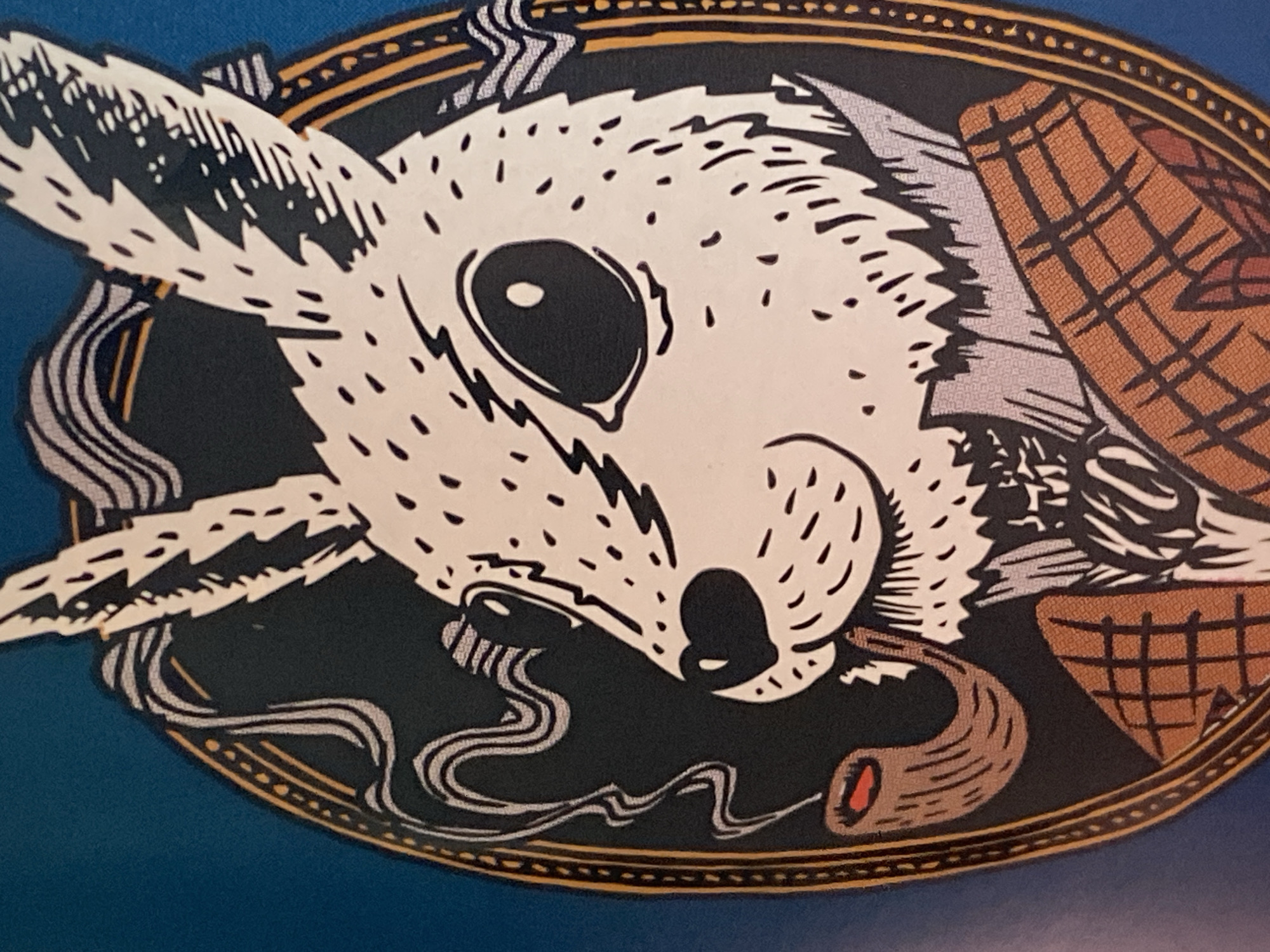 The image displays a stylized illustration of a white animal's head, most likely a rabbit or hare, rendered in a graphic, outlined style within an oval frame. The animal features large black eyes with a white highlight, a black nose, and white fur with numerous small black speckles. Its ears are prominent, extending upwards and backward.
The rabbit is depicted with anthropomorphic characteristics, evoking a detective or intellectual persona. A brown, checkered deerstalker hat rests on its head, partially visible behind one ear. A gray garment, possibly a scarf or cloak with wavy black stripes, is draped around its neck and shoulders. A reddish-brown pipe with a red tip is held near its mouth, emitting soft white curls of smoke, suggesting contemplation or relaxation.
The central illustration is enclosed within a distinctive oval frame, featuring a golden outer border with a dotted pattern and a solid black inner border. The background inside the oval is a dark blue-black, contrasting with the solid medium blue background outside the frame. The artwork lacks any visible text and does not provide specific cues regarding the time of day, weather, or a real-world location.