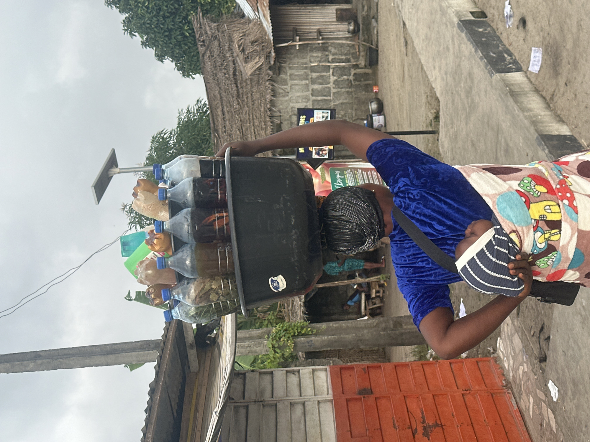 In the city of Badagry, Nigeria, a woman balances a large black basin filled with bottles of liquid and plastic bags on her head. She is carrying a baby wrapped in a colorful cloth with a mushroom pattern, secured to her chest. The woman wears a royal blue velvet top. She stands on a paved walkway next to a dirt road, with various buildings and vegetation visible in the background. To her left, there is an orange metal door and a wooden structure. Behind her, a thatched roof structure and brick buildings are apparent. The sky is overcast, suggesting a cloudy day. Scattered pieces of paper are visible on the ground.