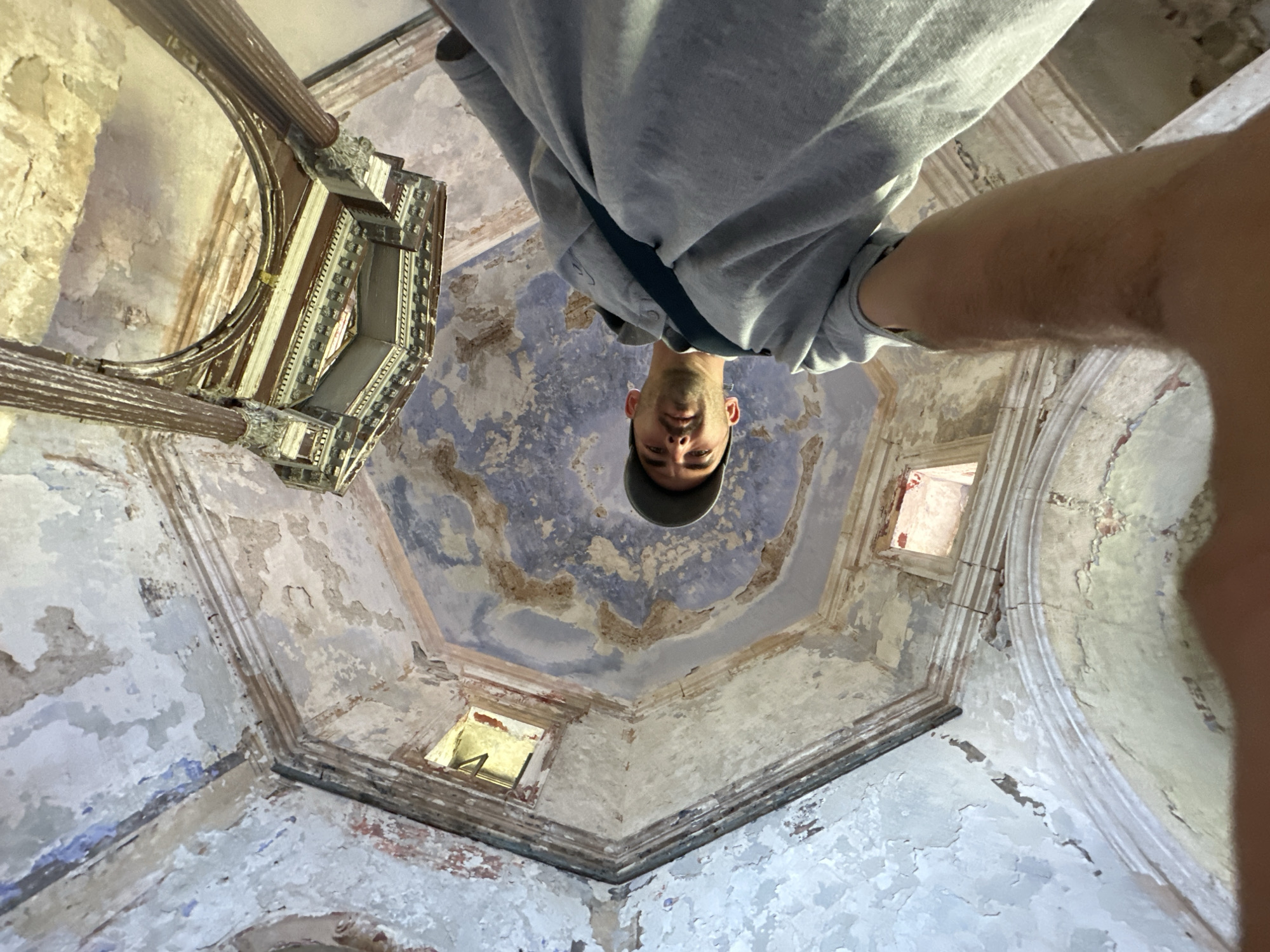 A low-angle, close-up shot shows a person wearing a gray hoodie and a dark baseball cap. The person's face is partially visible, looking upwards. The background reveals the interior of a dilapidated building with peeling paint and visible structural decay on the walls and ceiling. The architecture features an octagonal ceiling with decorative elements and rectangular openings that allow light to enter. In the upper portion of the image, there's a detailed view of an ornate wooden structure suspended from the ceiling, supported by fluted columns. The scene appears to be within the Church of Saint Nicholas in Prčanj, Montenegro, a location known for its historical significance and Baroque architecture, which has experienced periods of neglect.