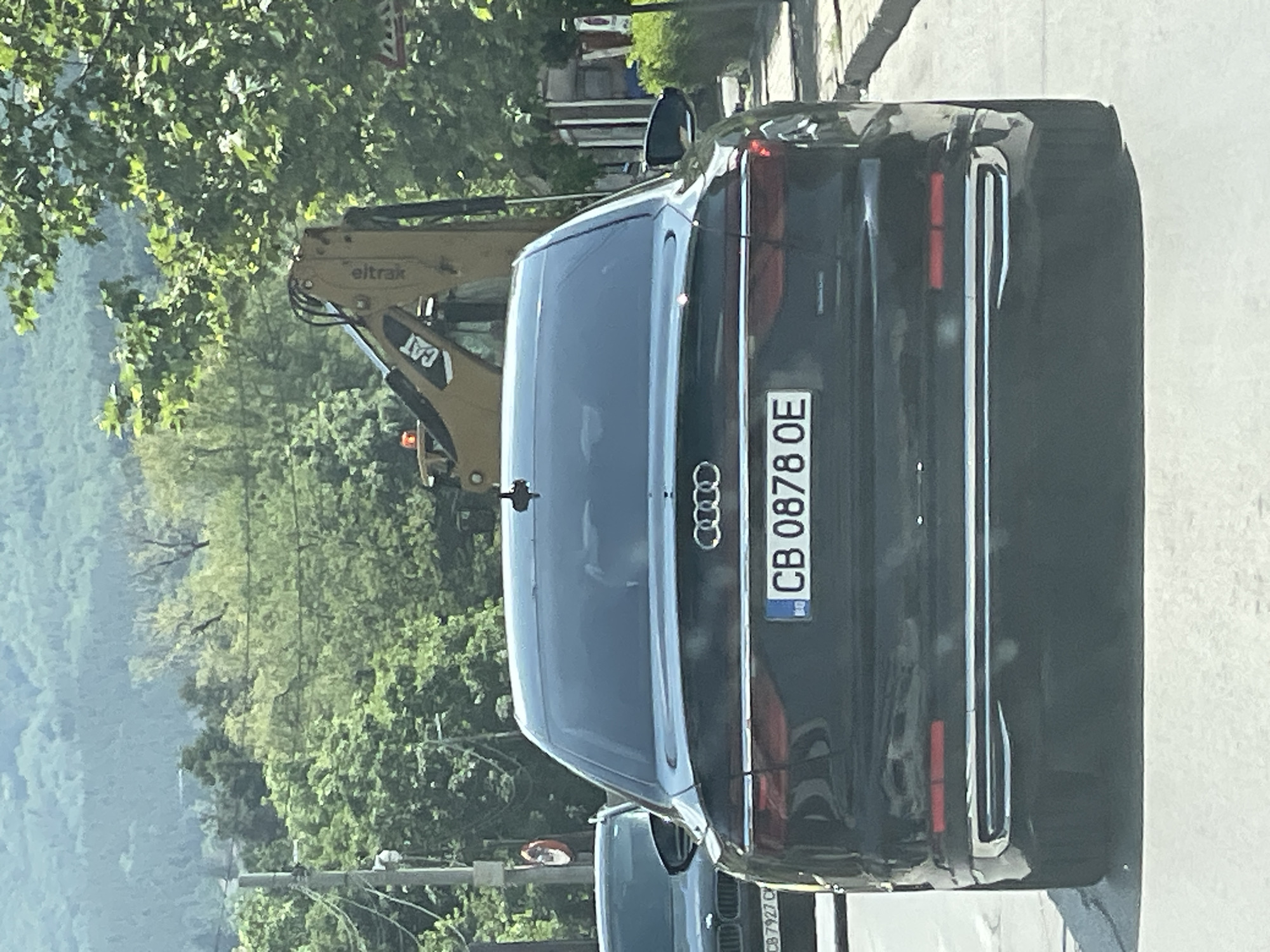 The image shows the rear of a black Audi sedan with a Bulgarian license plate reading "CB 0878 OE". The car is on a street, likely in Sofia, Bulgaria, given the license plate format. In the background, a yellow CAT excavator is visible, suggesting construction work. Trees and a hillside are also part of the environment, indicating an outdoor setting with some natural elements. Another car, a dark BMW with license plate "CB 7927 C", is partially visible to the left of the Audi. The lighting suggests it is daytime.
