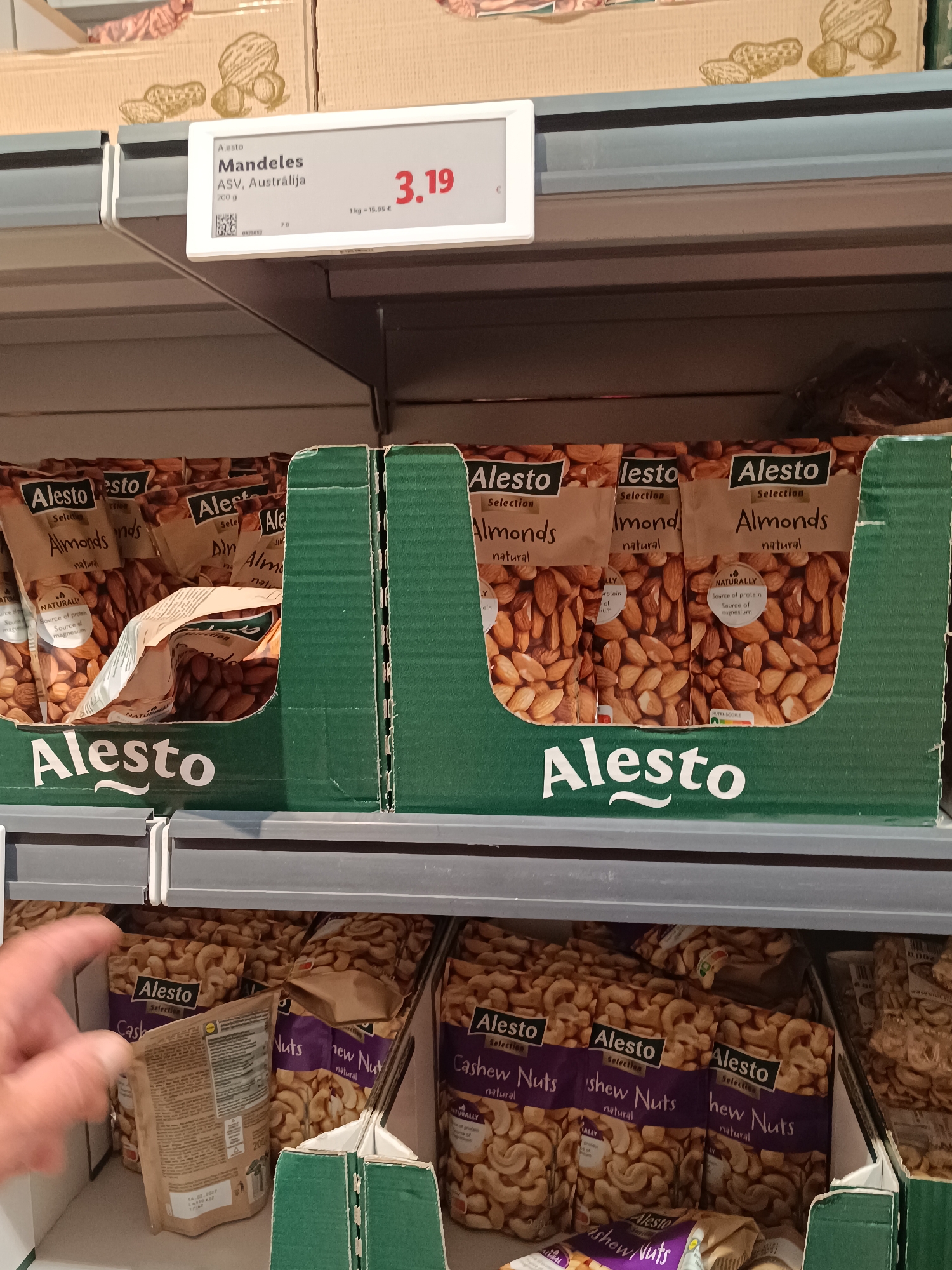 A store shelf display of "Alesto" brand nuts, primarily almonds and cashew nuts, is shown in Kuldīga, Latvia. In the foreground, a hand is reaching for a bag of cashew nuts on a lower shelf. The upper shelf displays a price tag for Alesto Mandeles (Almonds) from ASV, Australia, costing 3.19 Euros for 200 grams. The background shows other boxes of nuts and the store's shelving system. The lighting suggests an indoor retail environment.