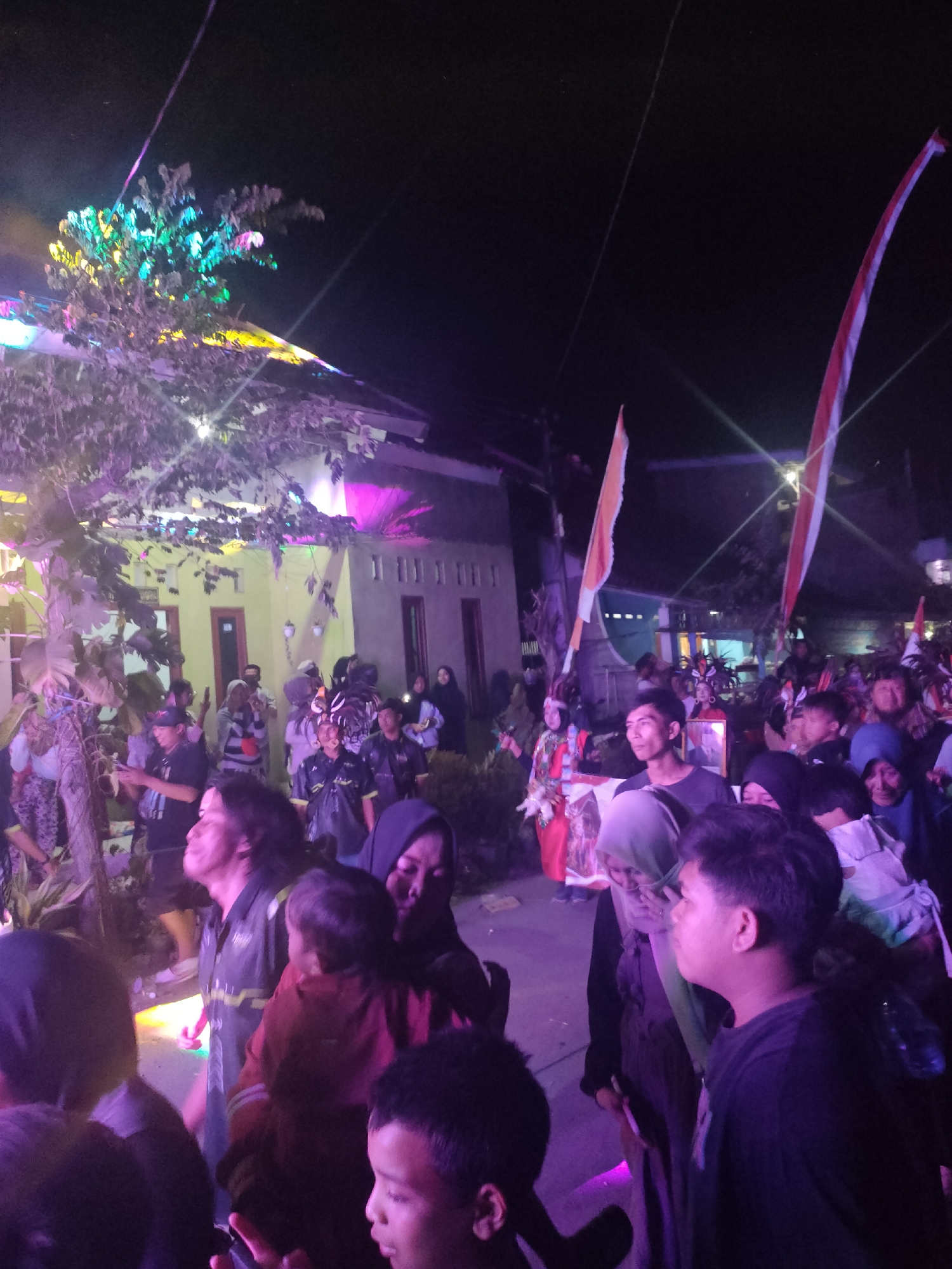This is a nighttime, eye-level, medium shot of a crowd gathered for an outdoor event, possibly a festival or celebration, in Tongaswetan, Indonesia. The scene is illuminated by a combination of colorful stage lights, including purple and green hues, and flag lights. In the background, a building with yellow walls and red-framed windows is visible. Two Indonesian flags, red and white, are prominently displayed, one on the left and one on the right side of the frame, each attached to a pole.
The crowd consists of people of various ages, with many individuals wearing casual clothing. Some are holding cell phones, likely to take pictures or videos of the event. The lighting casts long shadows and creates a vibrant, somewhat blurry atmosphere. The foliage of a tree, lit by the colorful lights, is visible in the foreground on the left, with its leaves appearing in shades of green and yellow. The overall mood appears to be festive and engaging, with people looking in various directions, some towards the perceived center of the event, others in conversation.