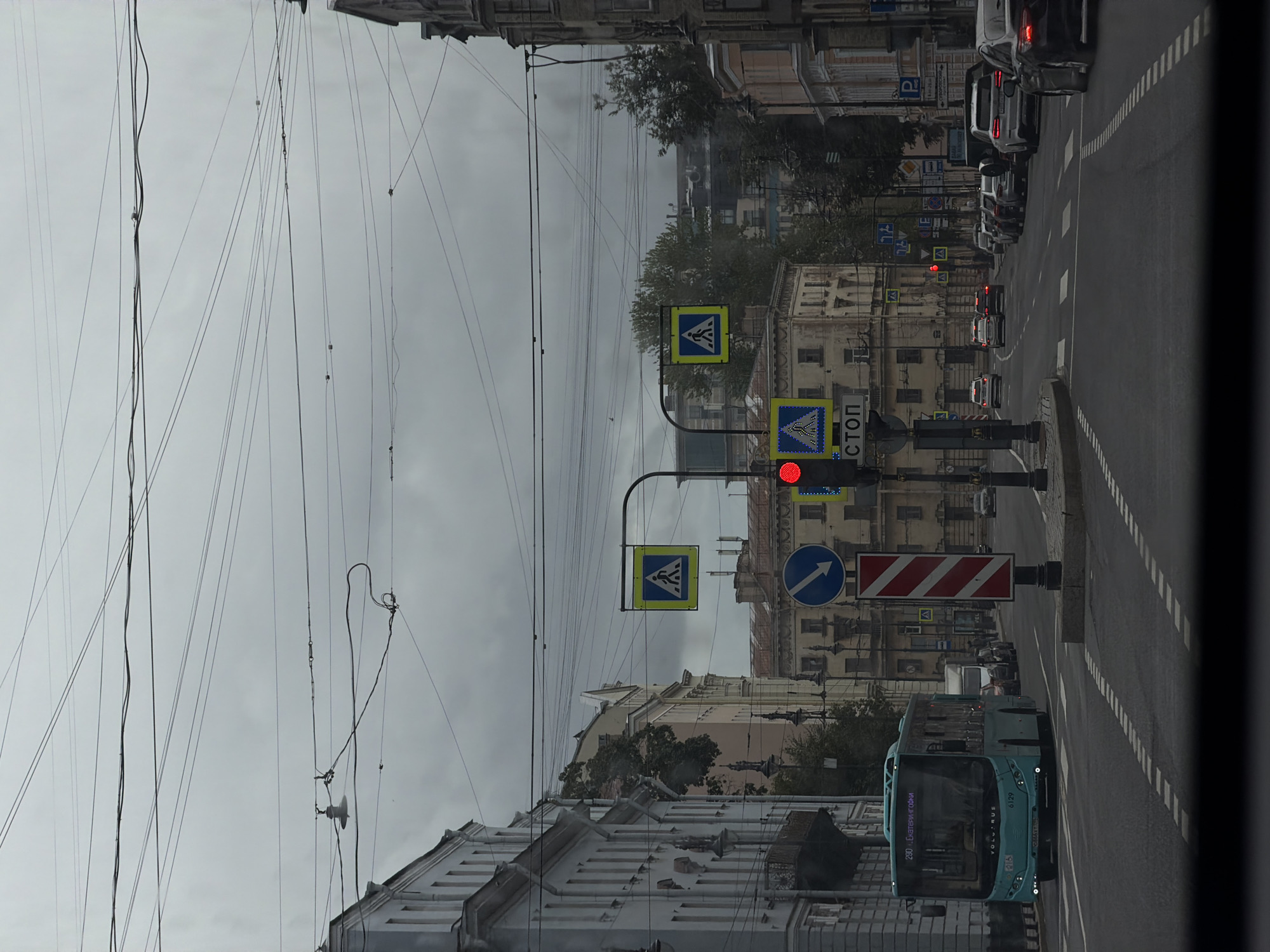 The image depicts a street scene in Saint Petersburg, Russia, on a cloudy day. A turquoise bus, with the number 290 and destination "Екатерингофки" displayed on its front, is driving down the road. The street is lined with multi-story buildings with ornate architecture on both sides. Overhead, a complex network of wires crisscrosses the sky, typical of a city with overhead tram or trolleybus lines. Traffic signals are visible, with one showing a red light. Several cars are also present on the road. Pedestrian crossing signs and directional signs are also visible. The road surface shows clear lane markings and crosswalks. The overall atmosphere is urban and subdued due to the overcast weather.
