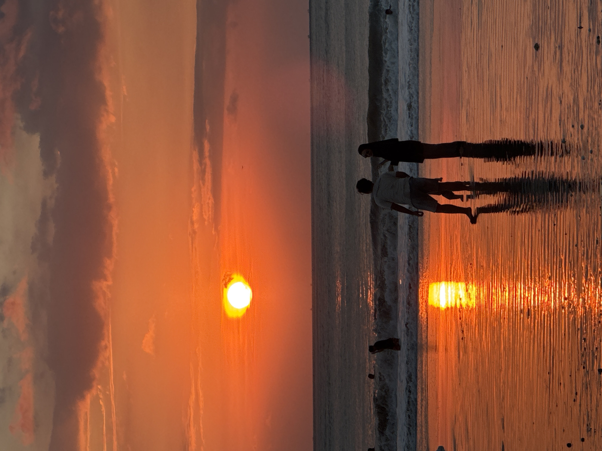 The image depicts a breathtaking sunset or sunrise over a beach, likely in Kuta Selatan, Indonesia. The sky is ablaze with vibrant hues of orange, yellow, and red, transitioning into darker, cloud-filled sections in the upper left. A bright, radiant sun is positioned low on the left horizon, casting a luminous glow across the entire scene.
Two figures stand in silhouette or near-silhouette by the water's edge on the right side of the frame. One taller person in dark attire stands slightly behind a shorter individual wearing a light-colored shirt and shorts, who appears to be walking in or interacting with the shallow water. Their long, stretched shadows are prominently cast across the reflective wet sand and water, extending towards the foreground. Gentle waves are visible breaking along the shore, with the frothy white lines catching the warm light. The sun's intense reflection creates a shimmering path of orange and yellow light on the water and wet sand, extending from the horizon towards the bottom right of the image. Other faint figures are discernible further along the beach in the distance. The overall scene exudes a serene and beautiful atmosphere, characteristic of a tropical beach during either dusk or dawn. There is no visible text in the image.