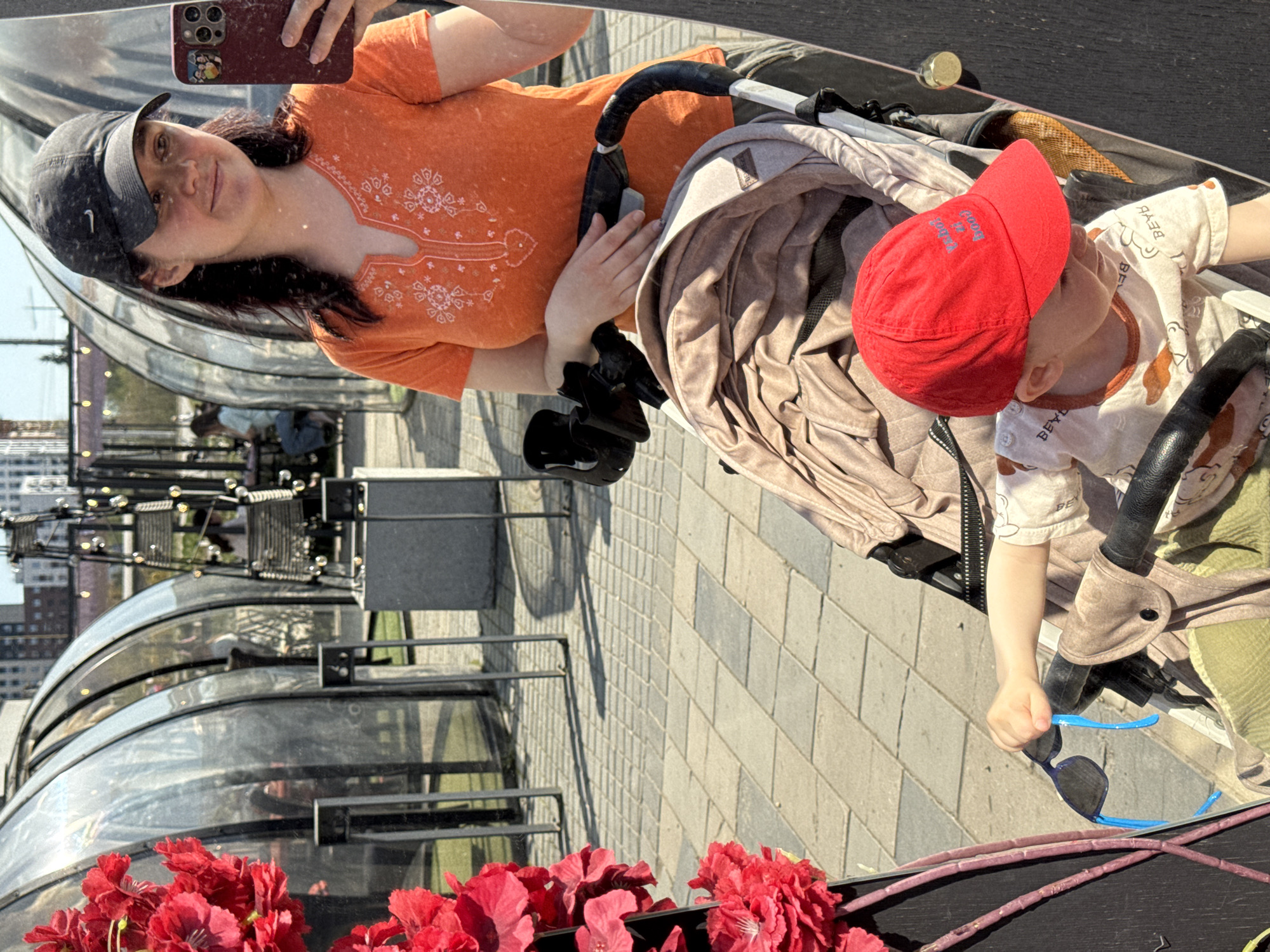 A reflection shows a woman in an orange shirt and a baseball cap taking a selfie. She is holding a mobile phone with multiple camera lenses. Beside her is a child in a stroller, wearing a red cap and a beige shirt with animal figures. The child is holding a pair of sunglasses. The background reveals an urban environment featuring a series of glass-domed structures. Red flowers are visible along the base of the reflection, suggesting they are part of the surrounding landscape. This photograph seems to have been taken in the city of Izhevsk, Russia.