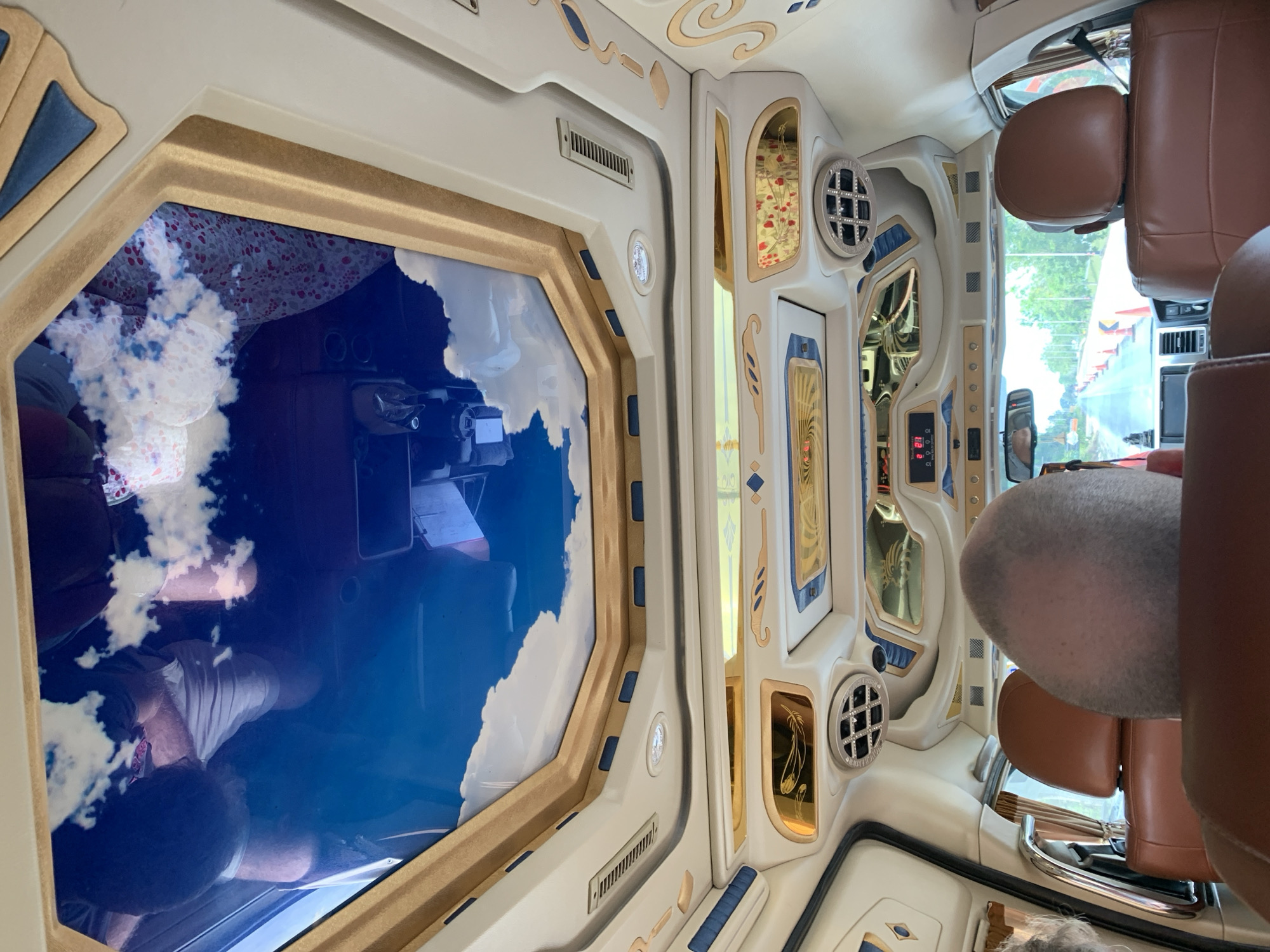 The interior of a vehicle, likely a van, is shown with ornate, custom designs in gold, white, and blue. Seated in the vehicle, a person with a shaved head is partially visible. In the background, there is a view of a road and trees, suggesting the vehicle is in motion. The image includes a reflection of people, objects, and a cloudy sky in a dark-tinted window. The general location is indicated as Tambon Thung Prang, Thailand.