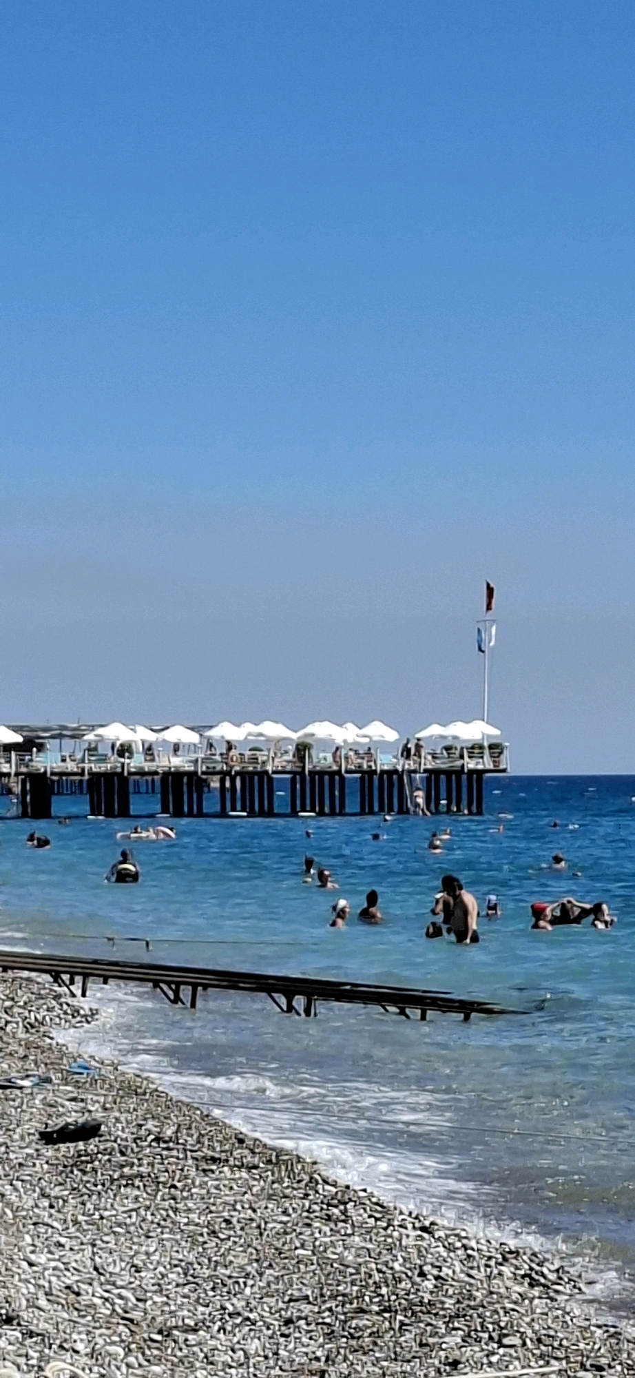 The image depicts a sunny day at a beach in Kemer, Turkey. A wooden pier with white umbrellas and people seated underneath extends over the clear turquoise water. Many people are swimming and relaxing in the sea. The shoreline is covered in pebbles. The sky is a bright, cloudless blue.