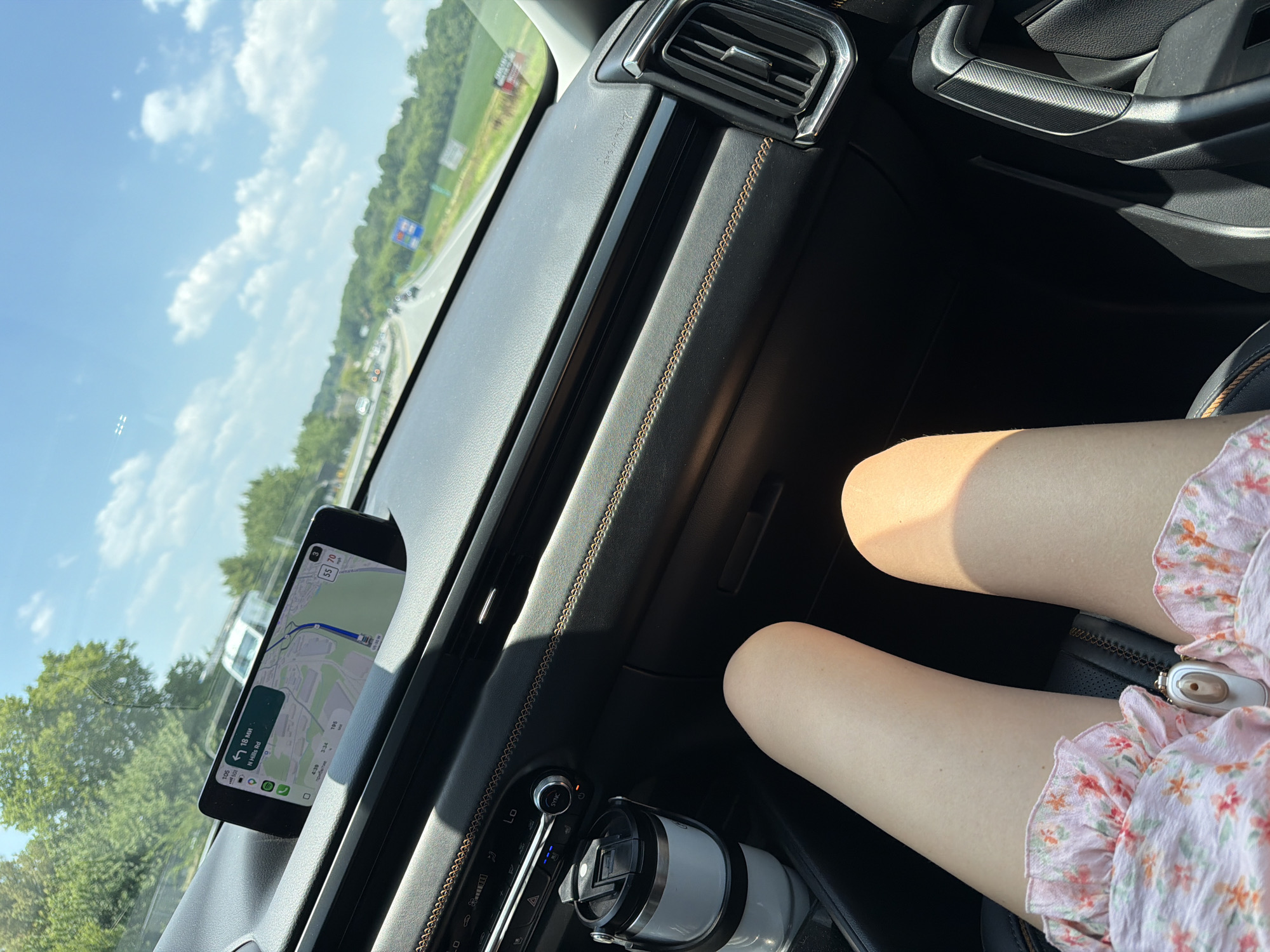This is a photograph taken from inside a vehicle, looking forward and slightly to the right. The perspective shows the dashboard, a smartphone mounted on the dashboard displaying a navigation app, a cup holder with a white tumbler, and a portion of the passenger's legs and dress. The exterior view through the windshield shows a highway with other vehicles, trees, and a bright blue sky with scattered clouds. Based on the navigation displayed on the phone, the location is East Hempfield Township, United States of America. The time shown on the phone is 4:38 PM. The speed limit indicated on the navigation is 55 mph.