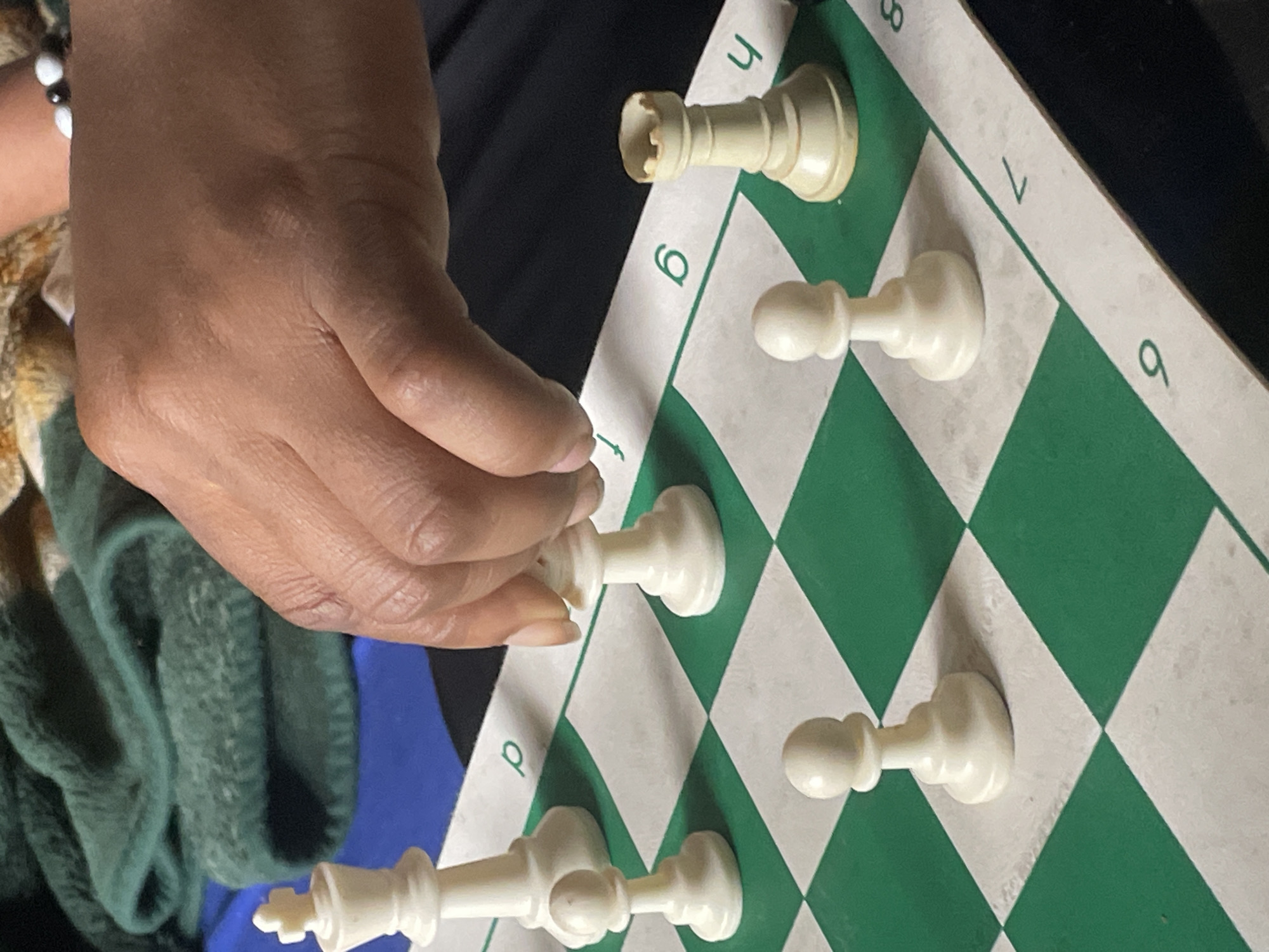 A chessboard with white pieces is oriented at an angle. A hand is shown reaching for a white piece on the chessboard. The chessboard features green and white squares with numbers and letters marking the rows and columns. The numbers 7 and 8 are visible. A white rook is placed on h8. A white pawn is on g7. A white pawn is on f6. A white pawn is on d4. A white king and queen are on the bottom row, left-hand side. A hand is on the chessboard, seemingly moving or about to move a white piece. The person is wearing a bracelet made of white and black beads. A green and gold sweater is visible in the bottom left corner. A person wearing a black shirt is behind the chessboard. No information in the image relates to Jos, Nigeria.