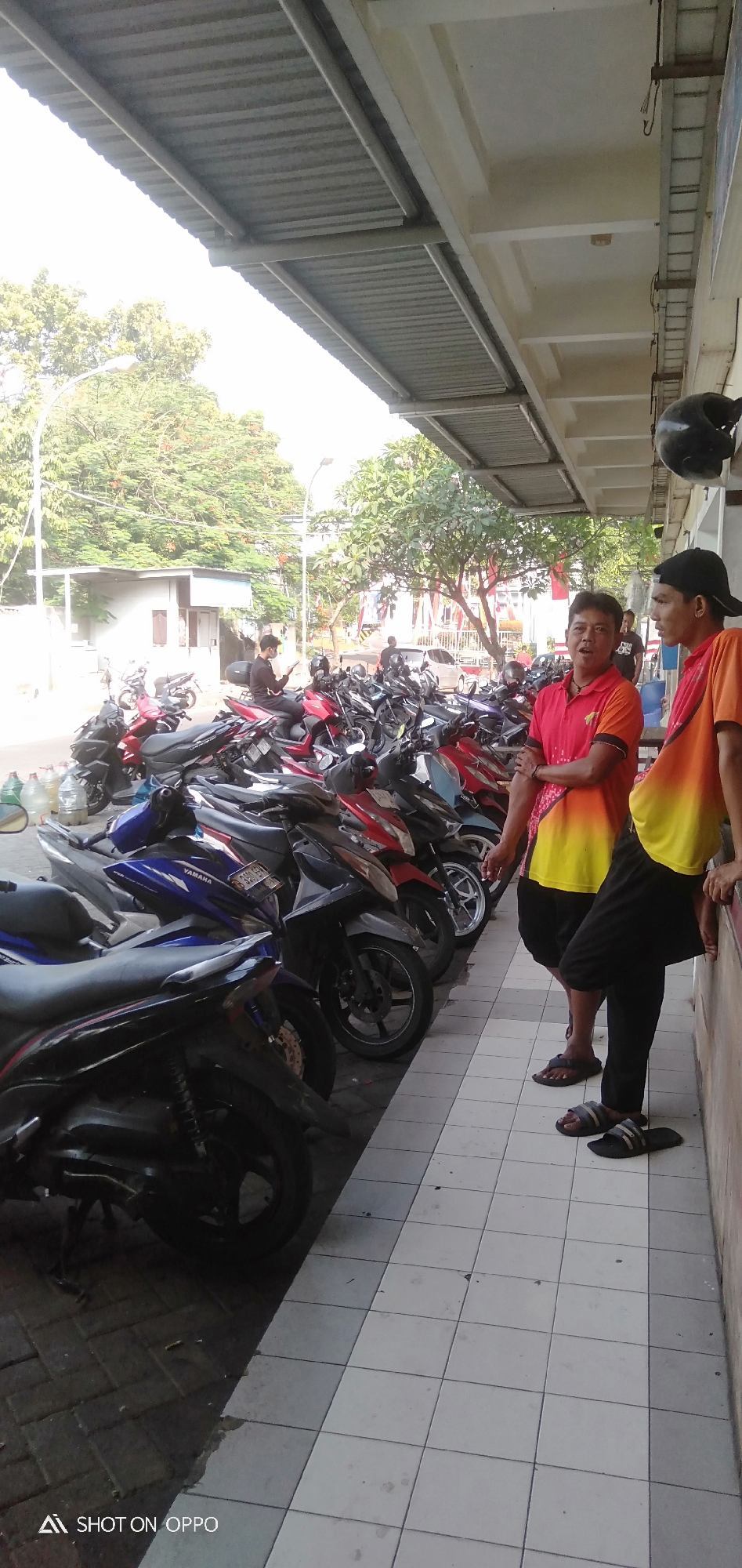 The image depicts a street scene in South Jakarta, Indonesia, with a prominent row of parked motorcycles. Two men are standing by the motorcycles, one wearing a red and yellow gradient shirt and black pants, and the other wearing an orange shirt and black pants. They appear to be casually conversing. In the background, more motorcycles are visible, along with trees, a building with a corrugated metal roof, and streetlights. The ground is paved with tiles. A black helmet is hanging on the edge of the building's overhang. The overall impression is of a typical street view in a city where motorcycles are a common mode of transportation.