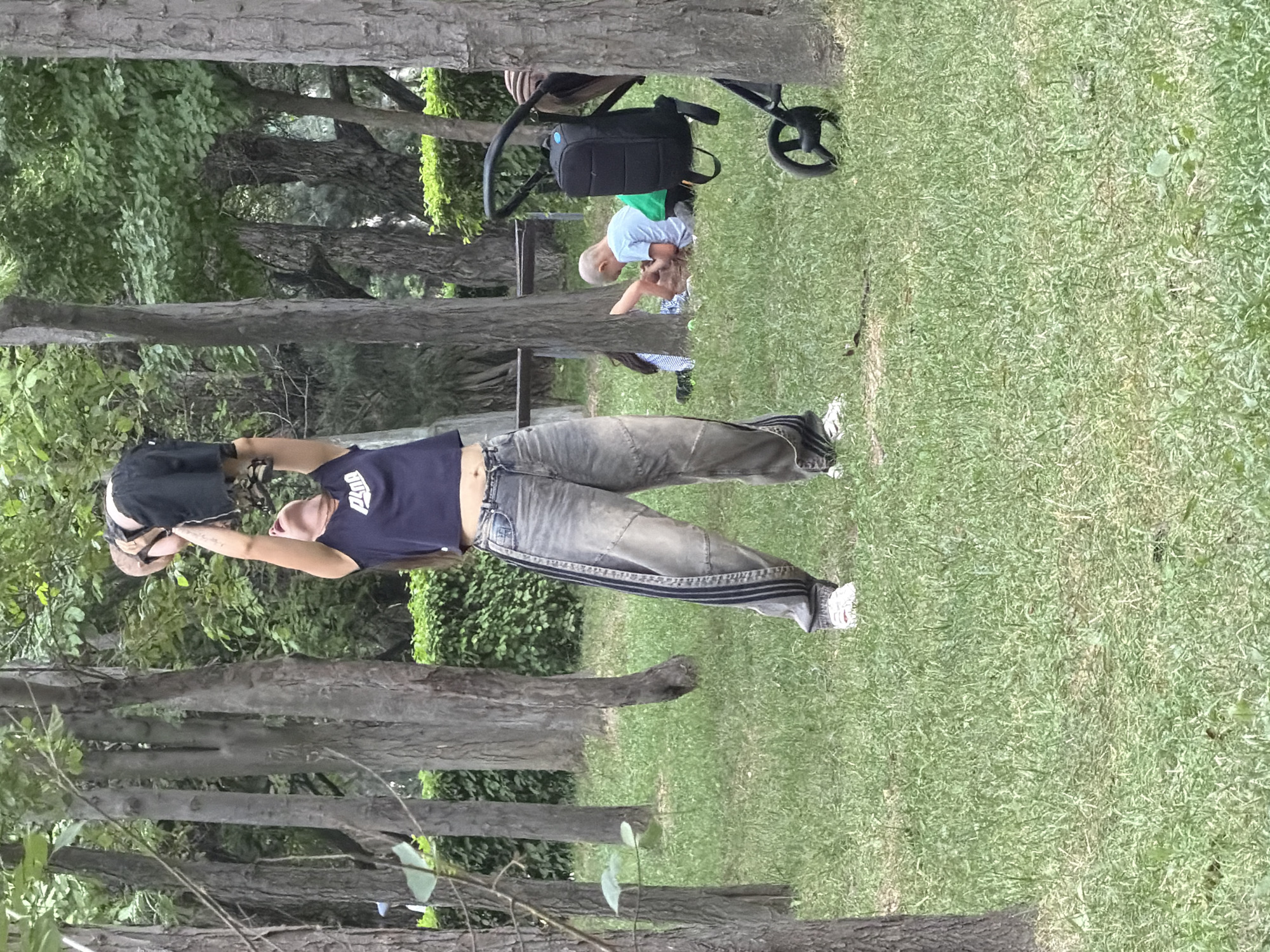 In a park, a woman stands in the center, holding a baby aloft. The woman is wearing a dark blue crop top with a white logo and baggy jeans with dark stripes on the sides. The baby, wearing black shorts and a light brown headband, is suspended over her head. To the right, a black stroller with a black backpack attached is parked on the grass. In the background, two children are lying on the grass, one wearing a light blue t-shirt and green shorts, and the other partially obscured. The environment is a grassy park with numerous trees and a dense green hedge in the background. The lighting suggests it is daytime.