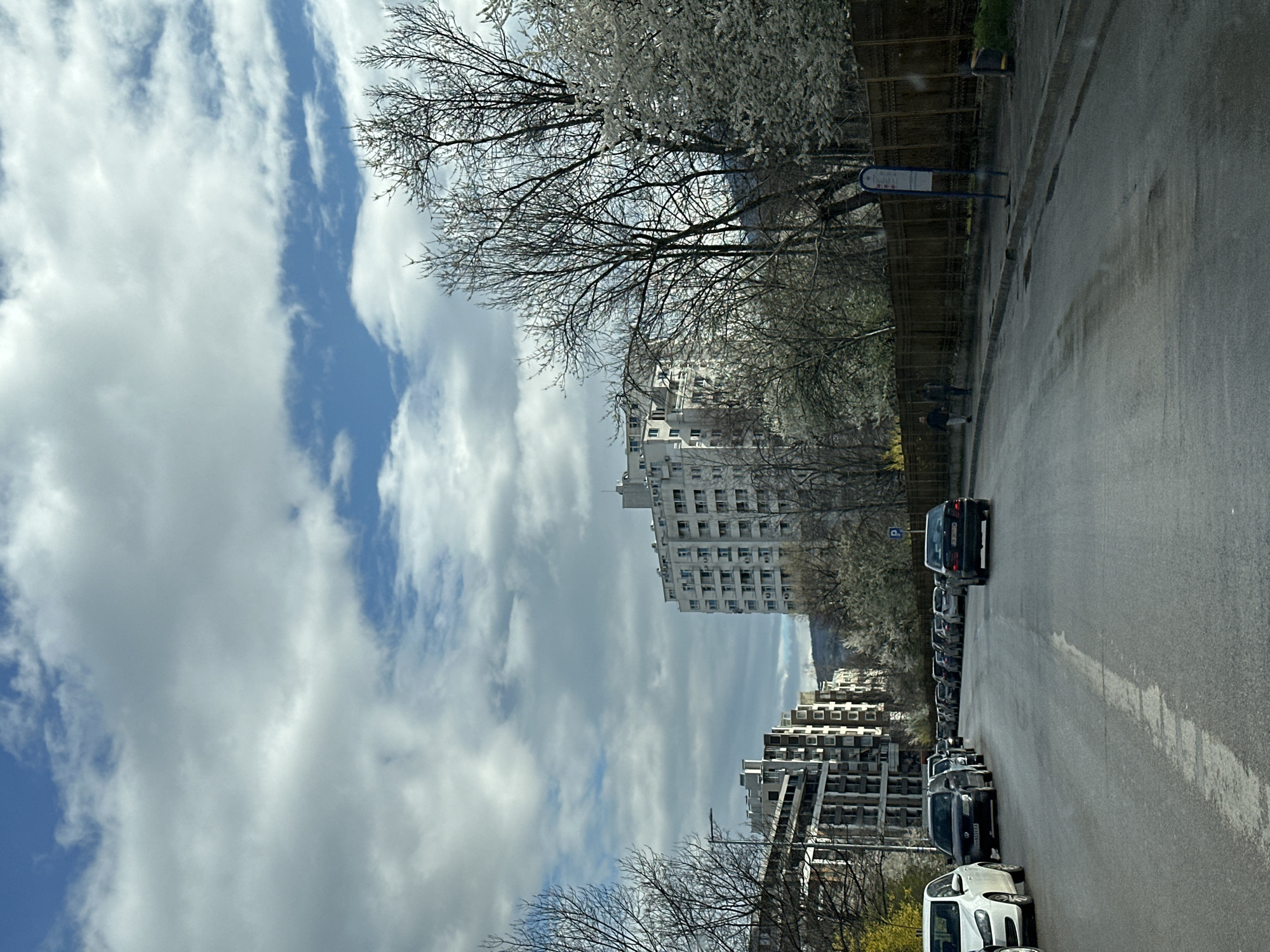 The image shows a street in Sofia, Bulgaria, lined with parked cars and buildings on either side. The sky is cloudy with patches of blue. Trees with budding or flowering branches are visible along the street. The road surface appears wet. A bus stop sign is on the right side of the street. People are walking in the distance on the right-hand side of the street.