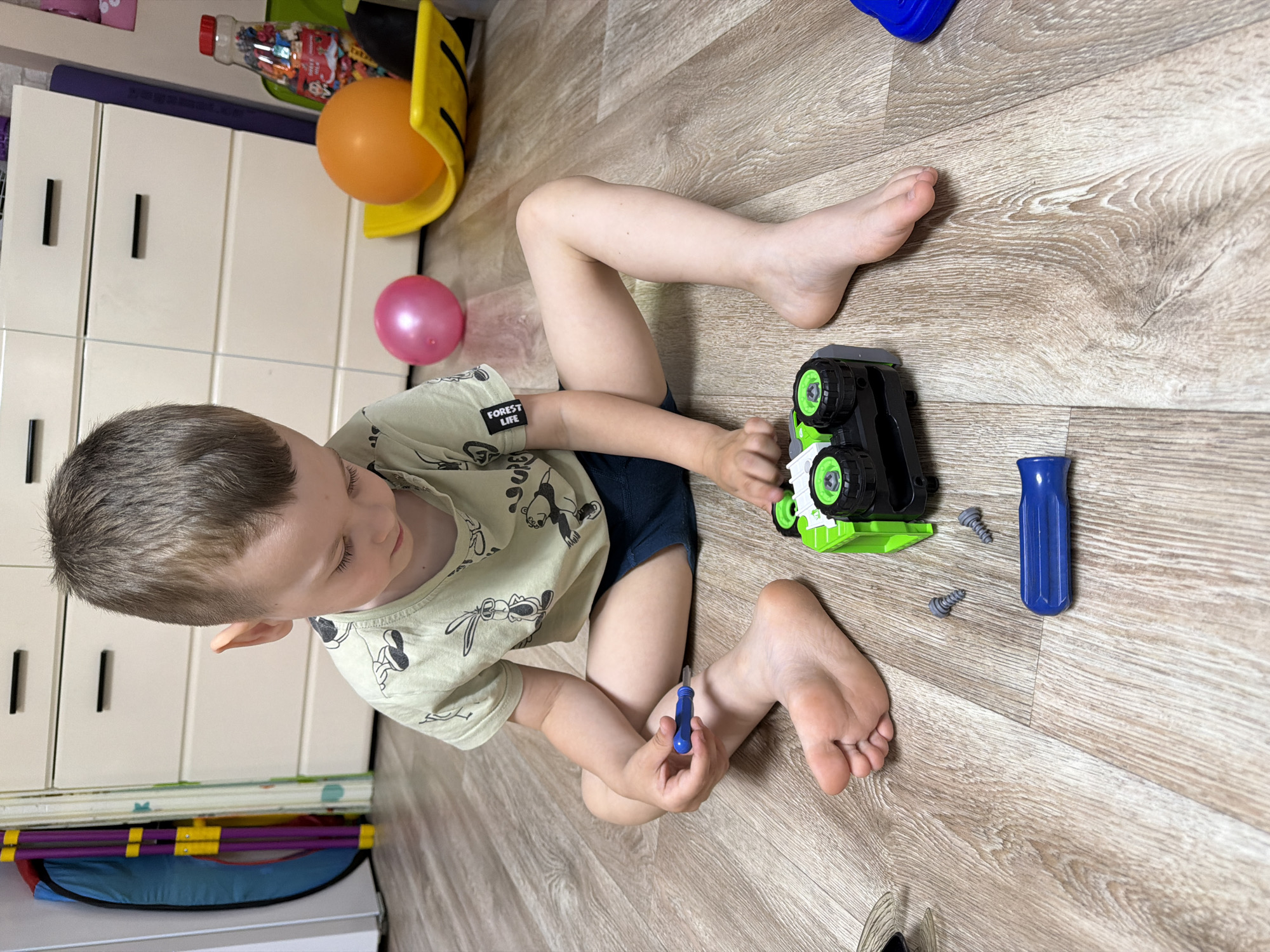 A young boy is playing on a wooden floor, likely in his home. He is lying on his back with his legs bent. In front of him is a green and black toy truck, and beside it are two screws and a blue screwdriver. The boy is wearing a light-colored t-shirt with a cartoon pattern. His eyes are closed, and he appears to be concentrating on his toys. The scene is brightly lit, suggesting it is daytime. In the background, a white dresser with drawers is visible, along with a yellow toy and an orange balloon. There is also a pink balloon nearby. The overall atmosphere is one of peaceful play. The t-shirt has the text "FOREST LIFE" on it.