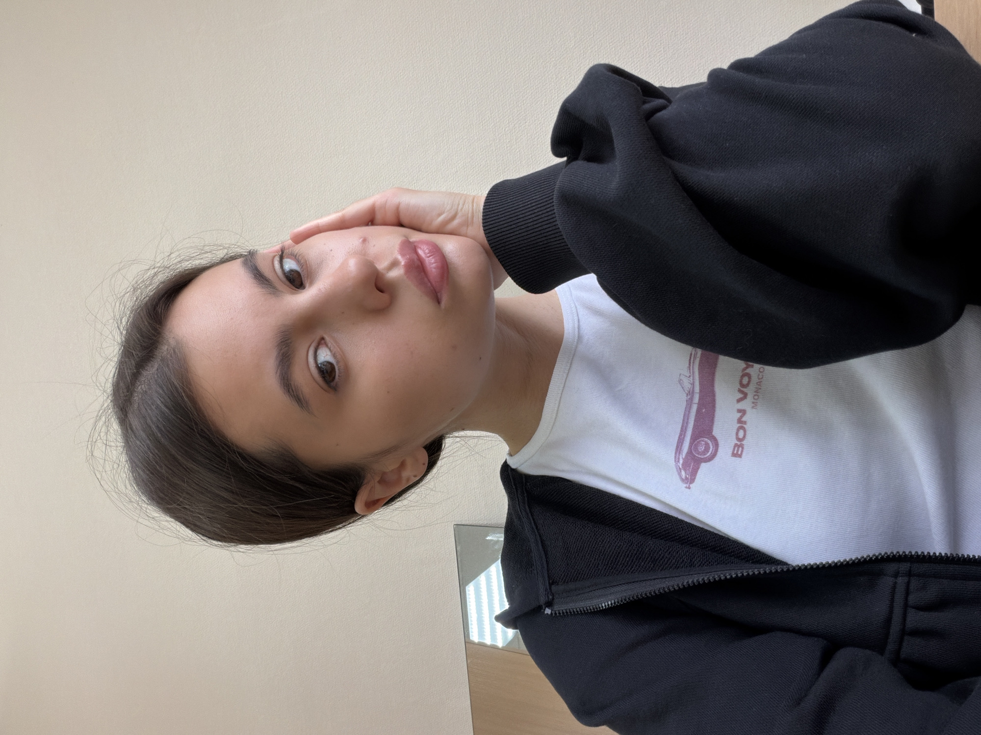 A close-up indoor portrait captures a young woman, likely a selfie, in Chaikovsky, Russia. She looks directly at the camera with a neutral to slightly contemplative expression, her dark eyes wide open. Her right hand is gently placed against her left cheek and jawline. She has dark hair, which is parted and visible around her face, and her lips appear to have a subtle, glossy finish.
She is wearing casual clothing: a black zippered hoodie, partially unzipped, over a white t-shirt. On the white t-shirt, a reddish-purple graphic of a vintage car, possibly a convertible, is visible. Below the car graphic, the text "BON VOY" and "MONACO" is printed.
The background consists of a plain, light beige or off-white wall. In the lower right of the frame, a reflection in a mirror or glass surface shows a glimpse of vertical light fixtures, indicating an indoor setting. The lighting is soft and even, characteristic of an interior environment.