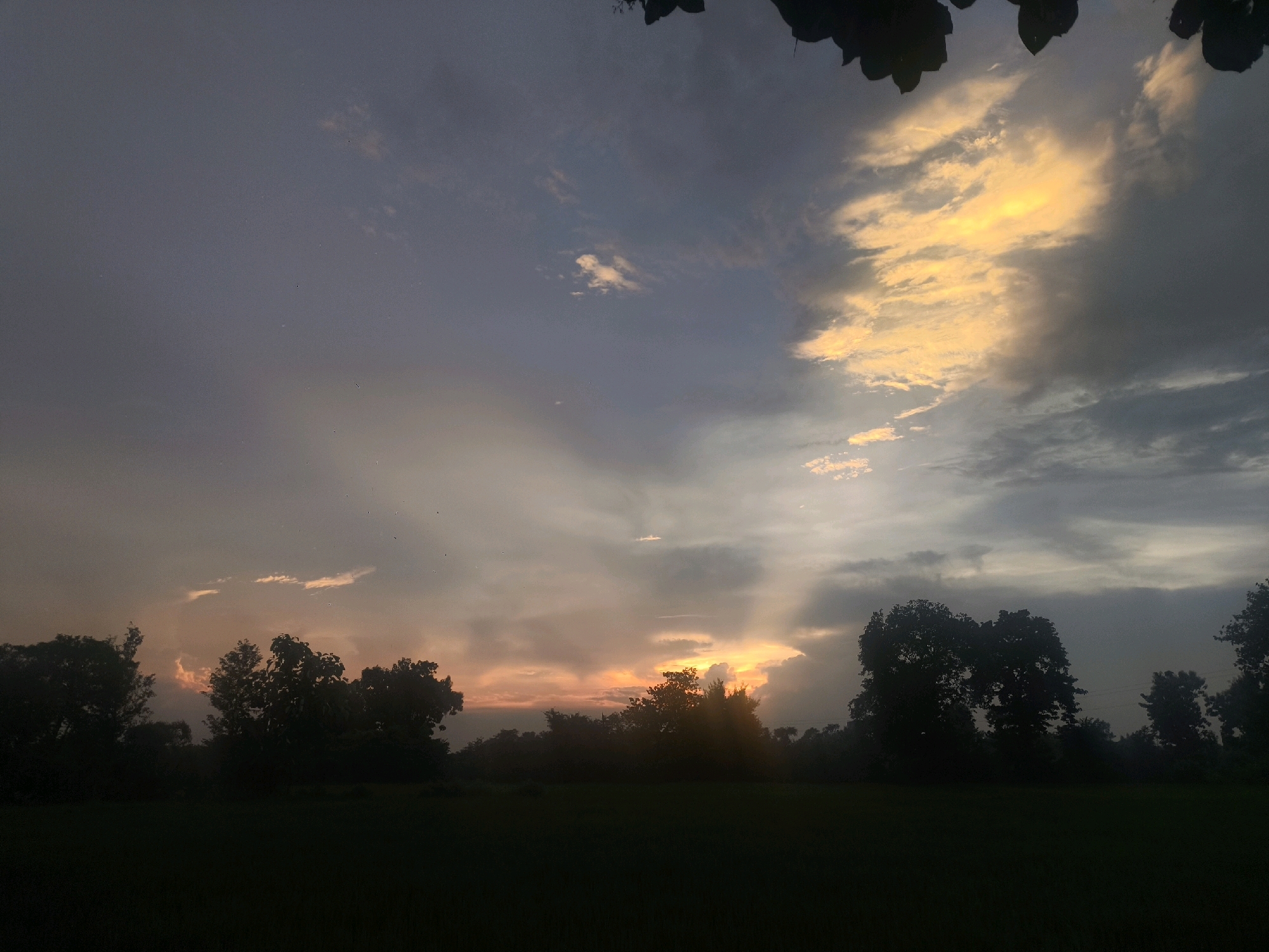 The image depicts a dusky landscape with trees silhouetted against a dramatic sky. The sky is filled with a mix of dark, heavy clouds and lighter, wispy formations that are illuminated by the setting sun. Shafts of golden light break through the clouds, creating a dramatic and atmospheric effect. The foreground is dominated by dark silhouettes of trees and what appears to be a field or open ground, rendered in deep shadow. The overall impression is one of a tranquil but somewhat moody evening in a rural or semi-rural setting, possibly in or near Kuinchwar, India, given the context. There are no visible people or animals.