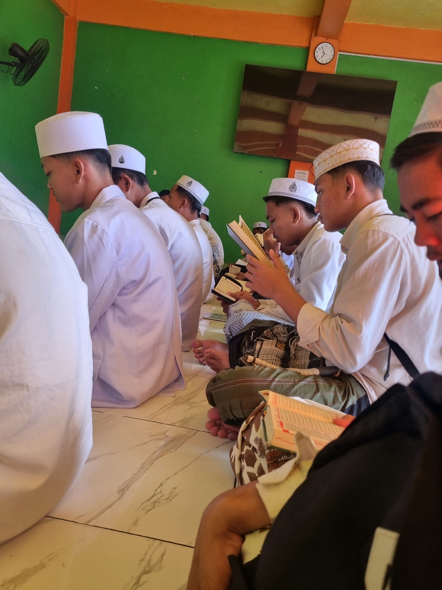 In an indoor setting with green walls and an orange ceiling, a group of young males are seated on a tiled floor, engaged in reading. They are all wearing white religious caps, commonly known as peci. The individuals appear to be participating in a religious activity, possibly a Quranic study session or prayer. One person in the foreground is holding an open book, likely a Quran, and their finger is pointing to a specific passage, indicating active reading or recitation. The lighting suggests daytime, with natural light possibly entering from an unseen window. A wall clock is visible on the upper wall, displaying the time. A ceiling fan is mounted on the left wall. The overall atmosphere is one of focused religious observance.