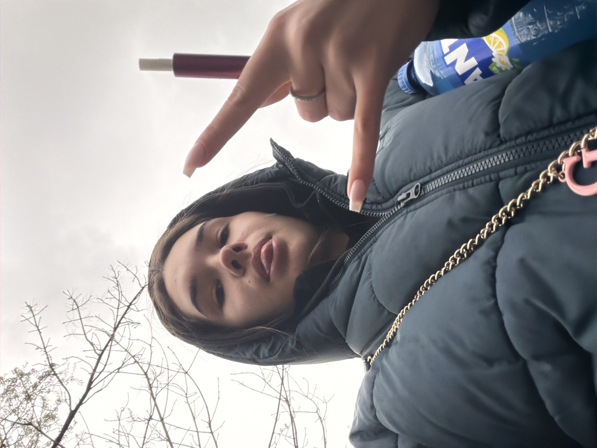 The image shows a young woman in a hooded, puffy, dark-colored jacket. A bottle with a blue label and a beverage inside is positioned behind her head, appearing to rest on her shoulder. A slender, dark object, possibly a cigarette or vape pen, extends from the upper-left of the frame. Her hand is in the foreground, fingers pointing downward. A gold chain is visible on the right side of the frame. The background features bare trees and a cloudy sky. Location cannot be verified.