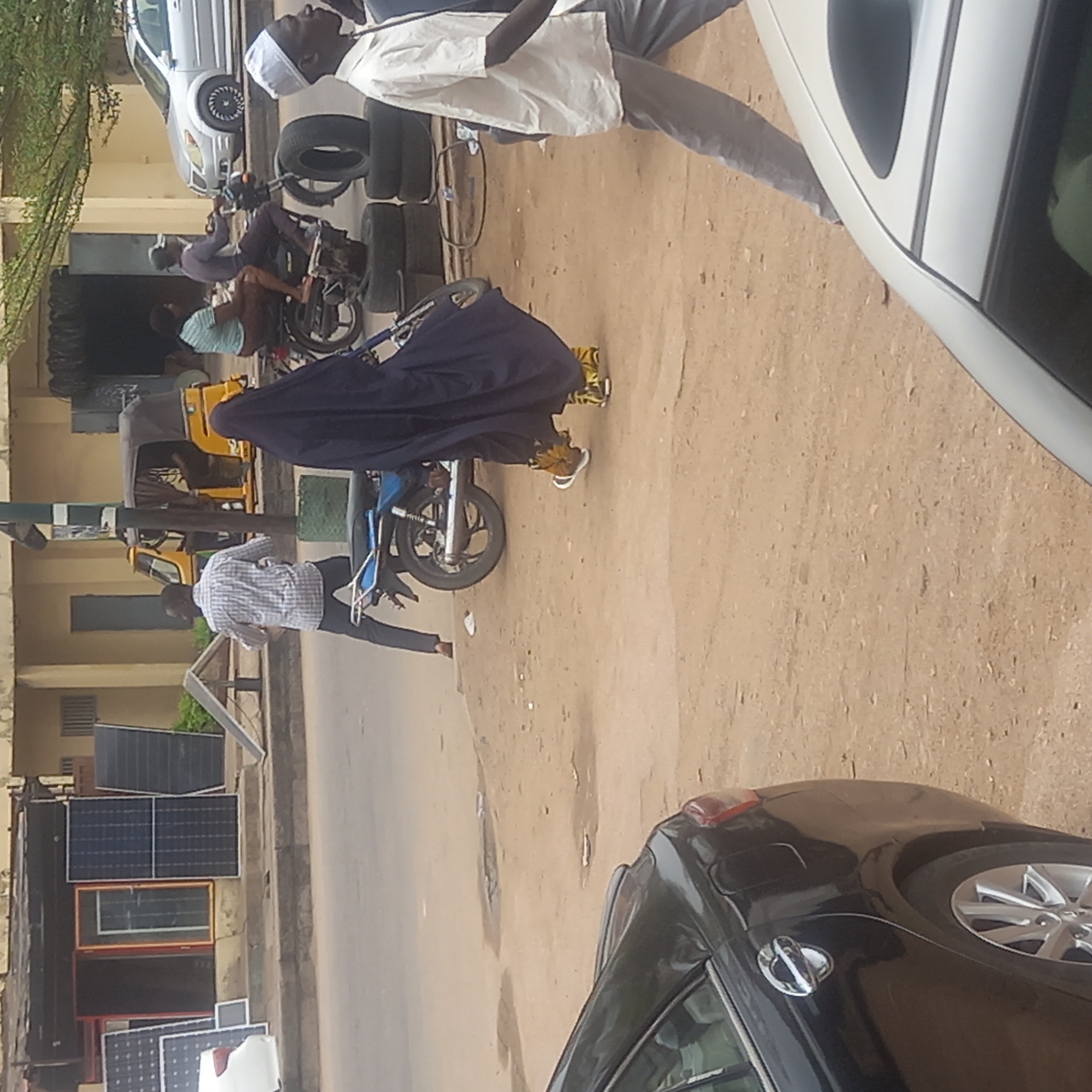 The scene is an outdoor urban environment in Dadi Riba, Nigeria. A sandy road is the central element. There are multiple vehicles present, including a black car in the foreground, a white car further back, a yellow auto-rickshaw (tricycle), and two motorcycles. Several people are visible. A man in a white shirt and a white cap is walking. A person wearing a full dark-colored covering is sitting on one of the motorcycles. Another man is standing nearby wearing a checkered shirt. Solar panels are stacked against a building on the left side of the frame. Used tires are stacked near the white car. Another man is working on a motorcycle near the stacked tires.