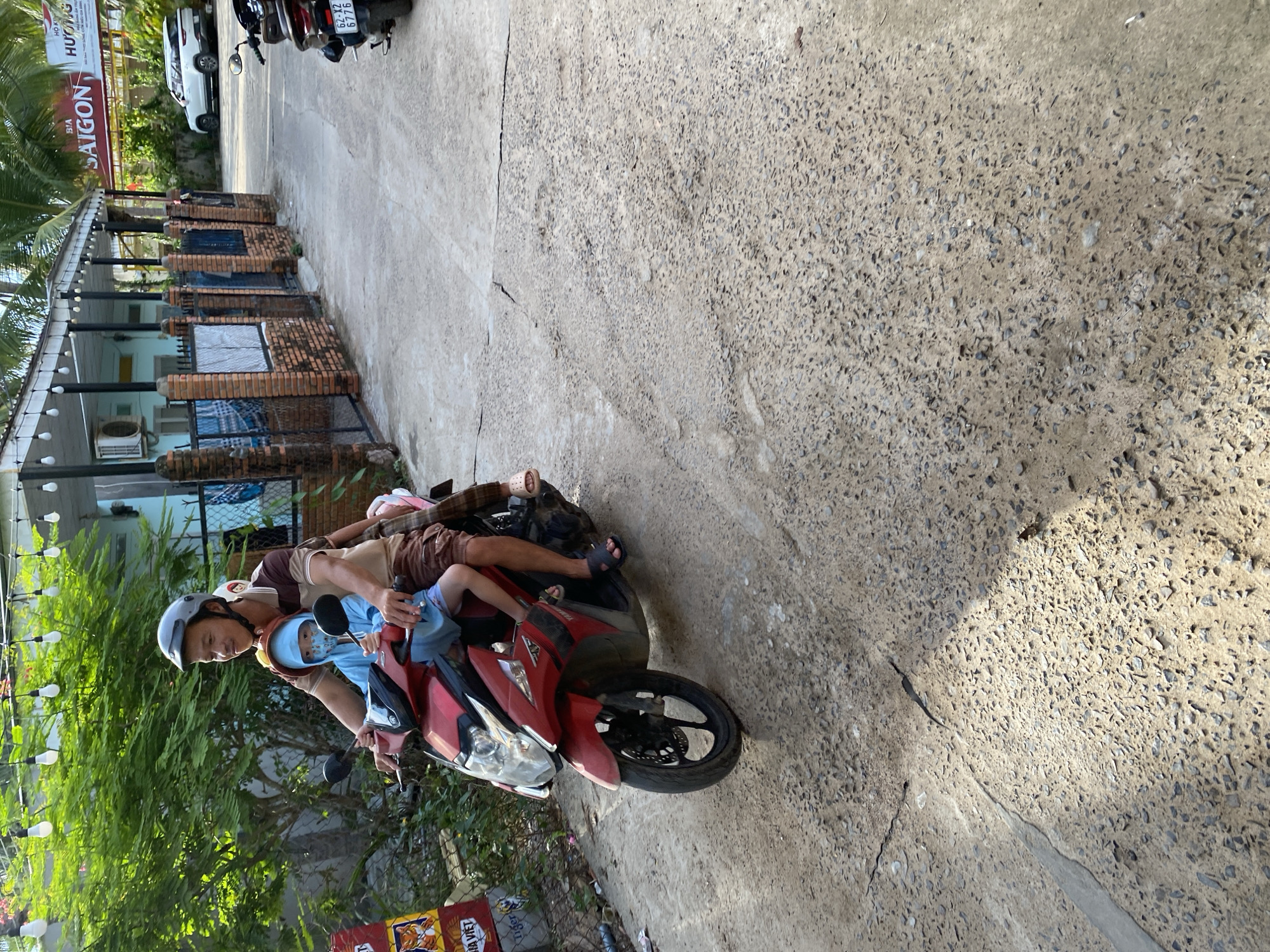 In Mũi Né, Vietnam, a man wearing a helmet and a patterned shirt drives a red Yamaha scooter on a paved road. A child wearing a blue hooded shirt and a face mask sits in front of him. The child has light skin and black hair. They are riding past a building with a blue facade and a brick fence. String lights with bare bulbs are strung above the building. A white SUV is parked on the right side of the road.