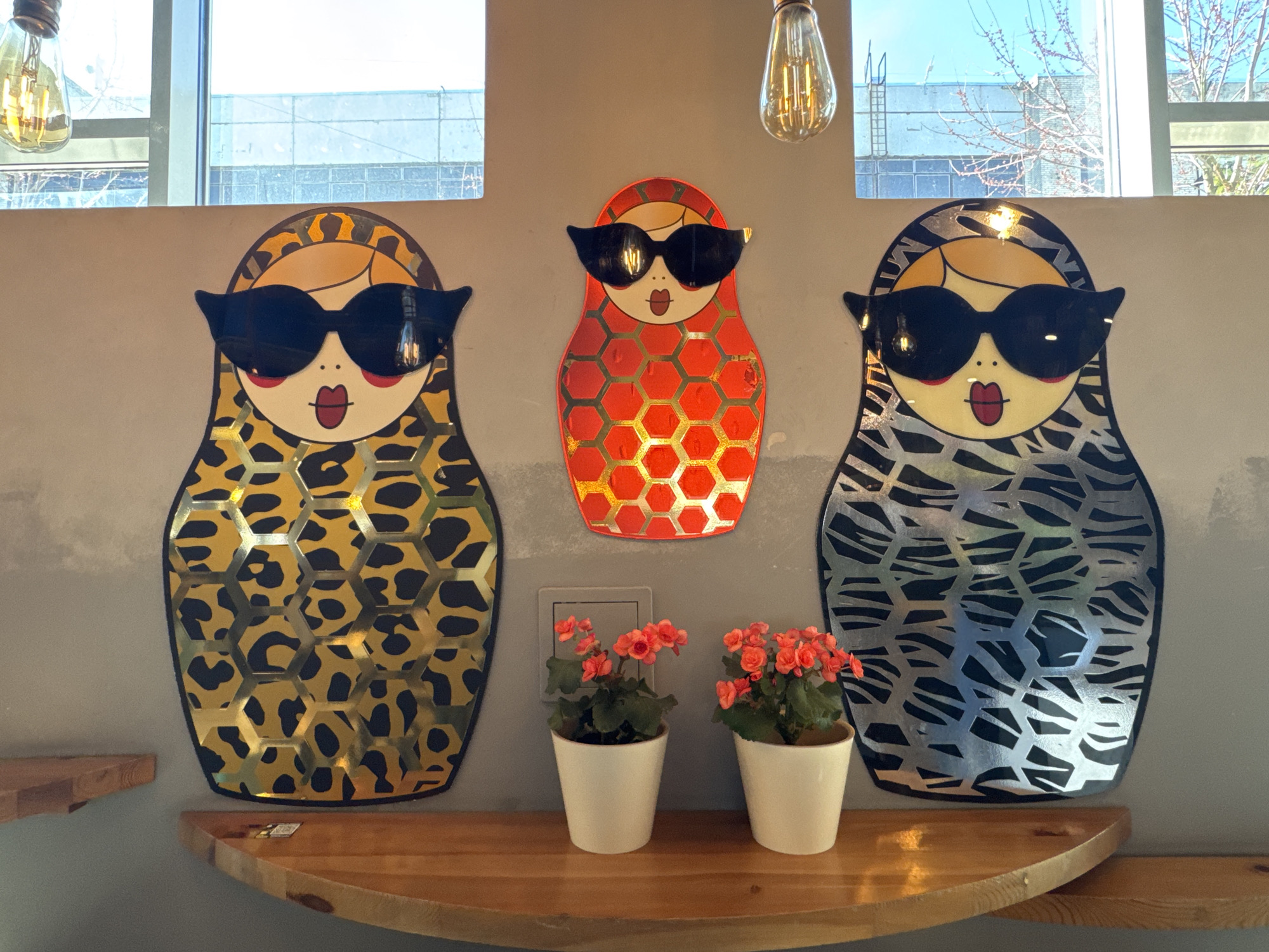The image displays a brightly lit interior wall featuring three stylized Matryoshka doll-shaped wall hangings and two potted plants on a wooden shelf. The wall is light gray.
The three decorative dolls depict feminine faces with blonde hair, prominent black cat-eye sunglasses, and red lips. The left doll features a leopard print body with a gold hexagonal overlay. The smaller central doll has a red body with a gold hexagonal overlay. The right doll displays a black and white zebra-like pattern with a silver hexagonal overlay. The surfaces of the dolls are reflective, catching light from the environment.
Below the dolls, a rounded, light-toned wooden shelf holds two small white potted plants, each bearing vibrant orange-red flowers. A power outlet is visible on the wall behind the plants.
Above the wall decor, two windows offer a view of the outdoors, revealing bare tree branches against a clear blue sky and distant buildings, indicating daytime. Two industrial-style Edison light bulbs with visible filaments hang from the ceiling, one on each side of the central window. The scene suggests a modern and decorative interior, possibly a cafe or stylish room, located in Saint Petersburg, Russia. There is no visible text in the image.