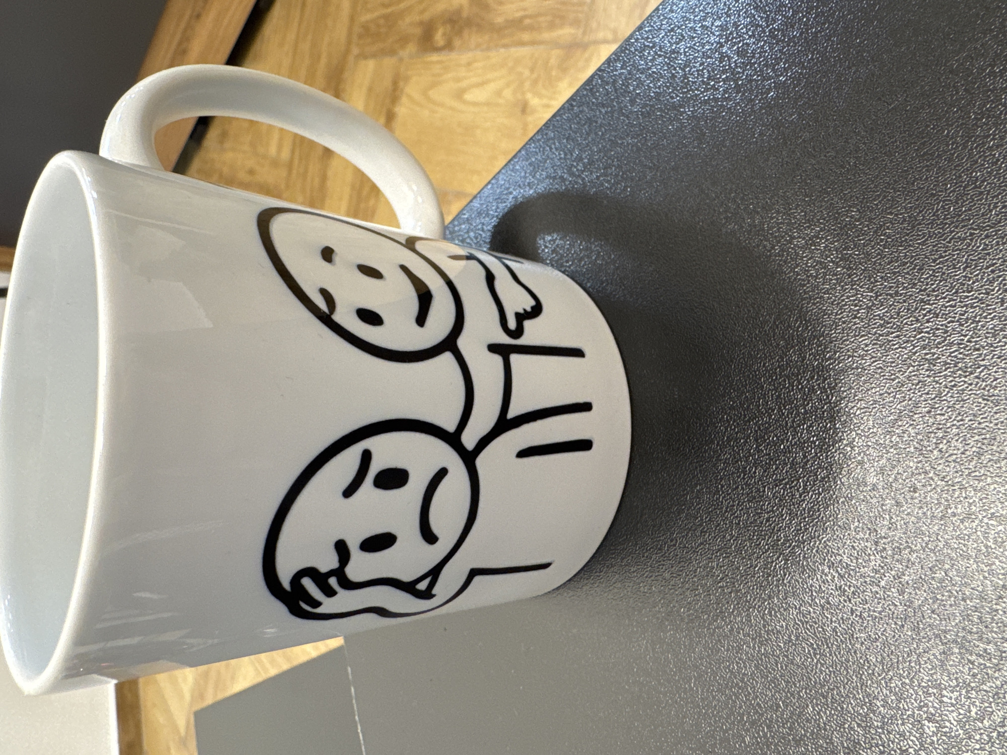 A white ceramic mug is depicted lying on its side on a dark, textured surface. The mug features black line art drawings of two stylized, stick-figure-like characters. The character positioned higher on the mug has a smiling, happy expression, while the character below it appears sad or unhappy, with a downturned mouth and its head seemingly resting on its hand. Both figures are connected, forming a singular, elongated body with two distinct heads.
The dark surface beneath the mug has a fine, pebbled texture. A soft shadow of the mug is cast on this surface, indicating an unseen light source. In the background, a warm-toned wooden surface with a visible grain is partially visible. The setting appears to be an indoor environment. The mug itself is glossy and clean, with its handle curving upwards in its current orientation. No discernible text is present on the mug; only the illustrative figures are visible.