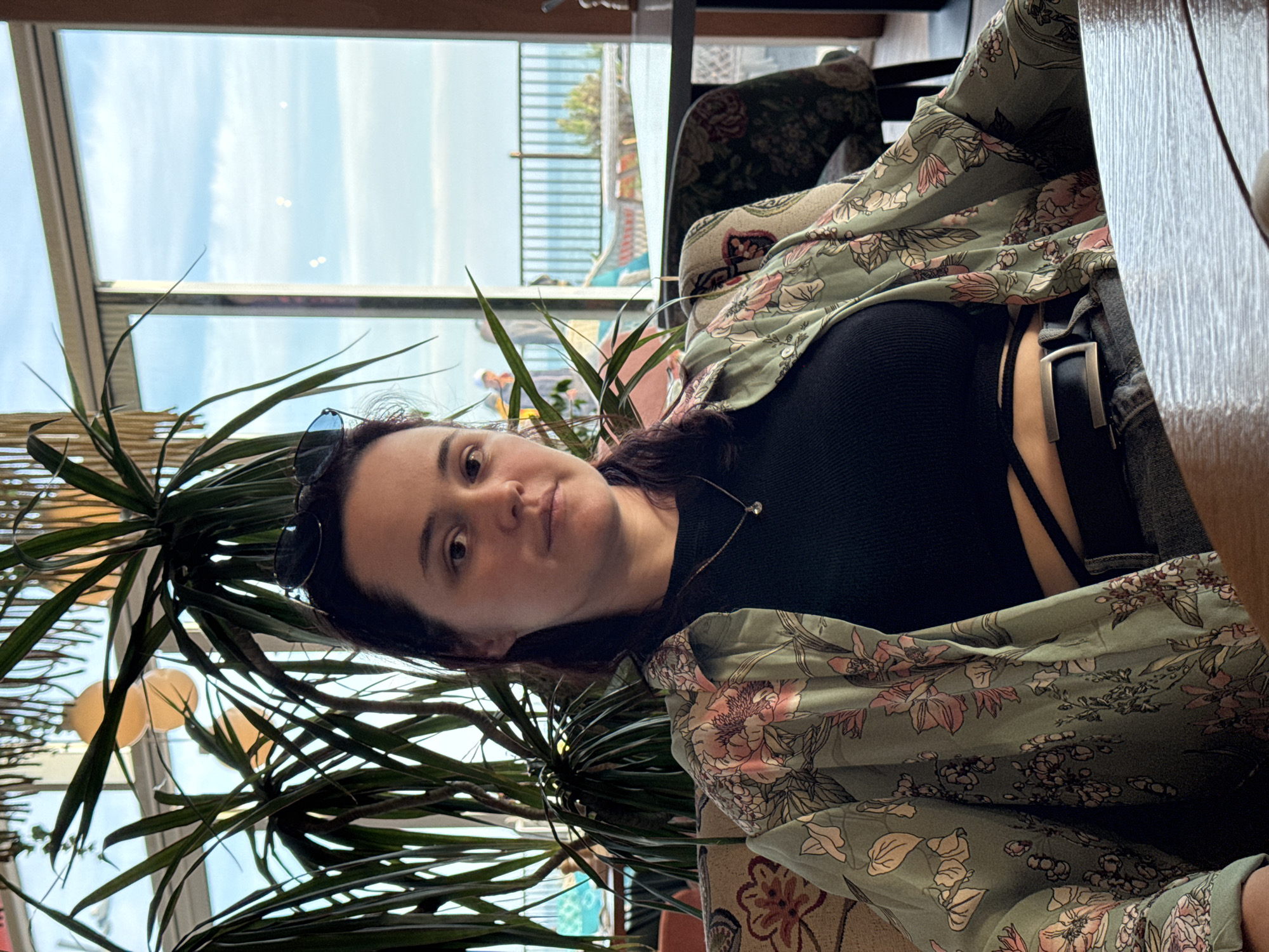 The image shows a woman with dark hair and sunglasses on her head, wearing a black top, a green floral-patterned kimono, and jeans with a black belt. She is sitting indoors, likely in a cafe or restaurant, as suggested by the chair she's sitting on and a table partially visible in the upper right corner. A large potted plant with long, slender leaves is prominent in the foreground. The background shows a bright, cloudy sky and a waterfront area with railings, indicating a coastal location. Based on the prompt, this location is in Sochi, Russia.