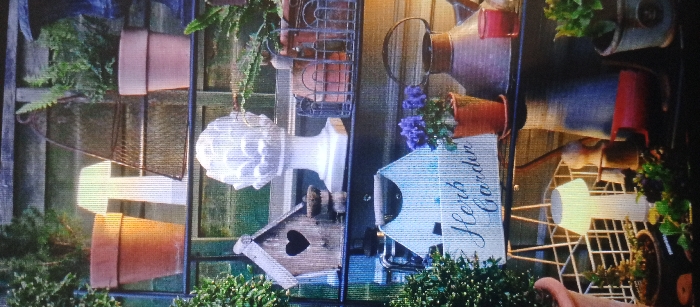 The image displays a vertical composition, rotated 90 degrees clockwise, depicting a static arrangement of gardening-themed decorative items and plants against a wooden background in the city of Unknown, Unknown. No individuals are present, and no actions or interactions are occurring.
The scene features a multi-tiered display unit or shelves. In the upper-left quadrant, green foliage, a wooden fence panel, and an unglazed terracotta pot containing a plant are visible. Below this, another terracotta pot, a metallic mesh object hanging, and a prominent white decorative finial with a fluted design are arranged. Directly beneath the finial is a small wooden birdhouse featuring a heart-shaped entrance. The lower-left portion shows dense green foliage.
In the upper-right quadrant, a rustic, aged metal watering can is positioned above a small terracotta pot containing purple flowering plants. Adjacent to these is a rectangular white sign inscribed with "Herb Garden" in a cursive font. The mid-right section includes a white open-lattice stand, various terracotta pots (some empty, some with small green plants), and a reddish-brown watering can. The lower-right corner reveals additional green foliage and the rim of an obscured terracotta pot. The background consists of vertical wooden panels, possibly part of a fence or wall, in muted green and brown tones. The lighting is uniform, casting soft shadows.