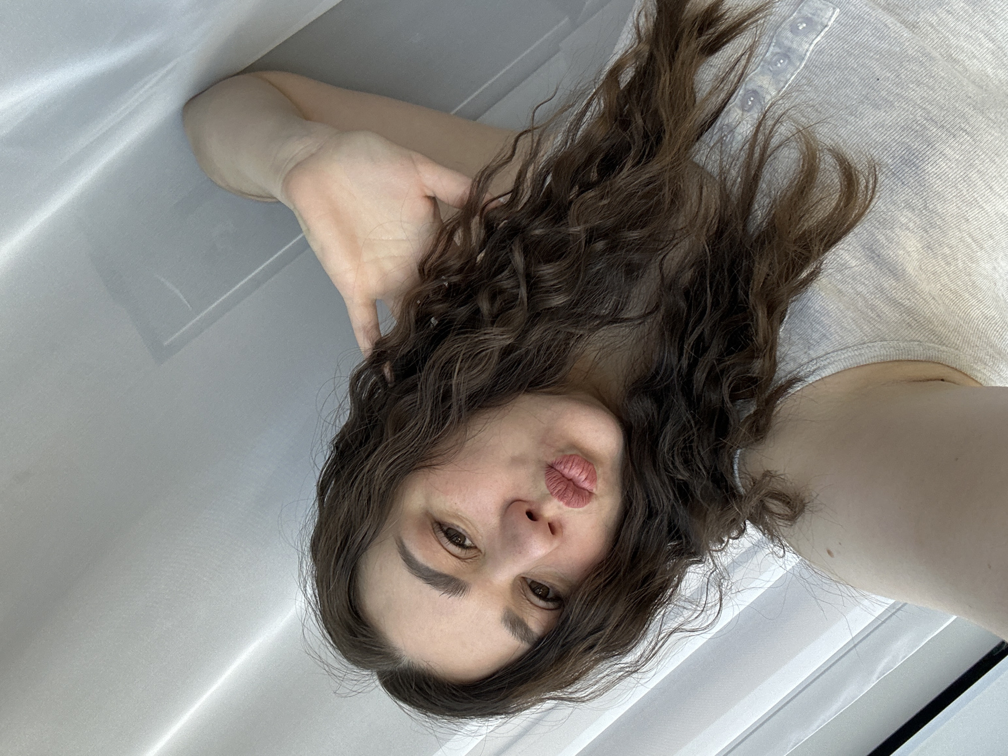 A woman with brown curly hair and fair skin is pictured from a close, tilted perspective. She is lying down, with her head near a white, vertically-oriented object that appears to be a curtain or blind. The woman is pursing her lips, and her left hand is positioned near her head. She is wearing a light-colored top with button details. The location is not discernible from the image. No details about Chaikovsky, Russia are discernable from the image.