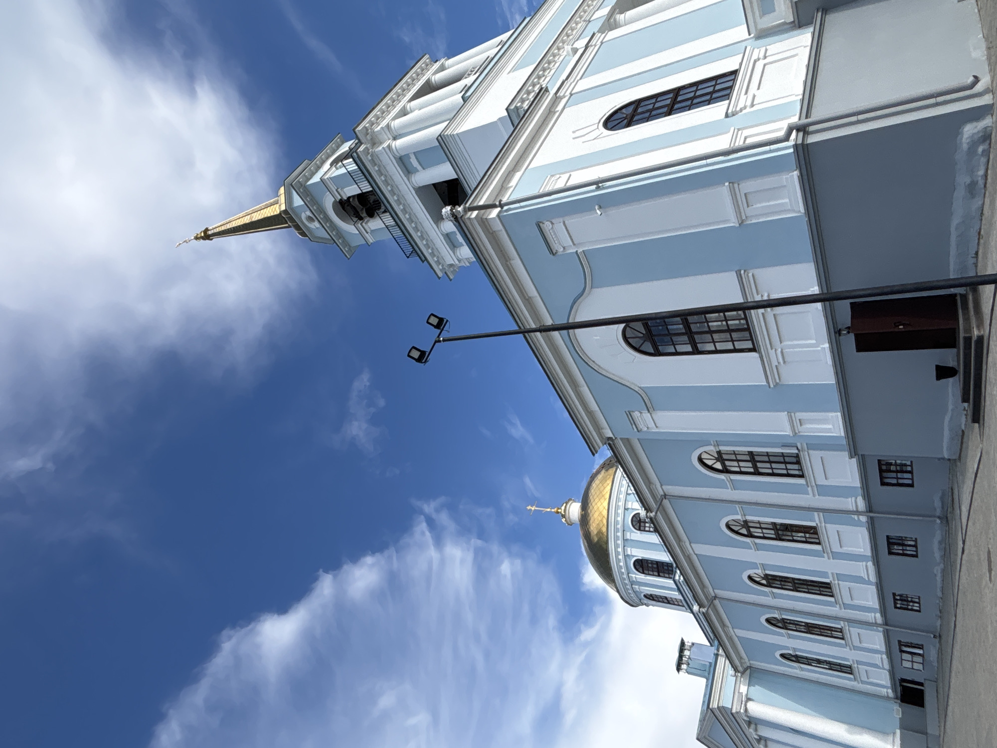 The image shows a low-angle view of a blue and white church with a golden dome and a tall spire against a bright blue sky with scattered clouds. The church has multiple arched windows with dark frames and classical architectural elements like columns and decorative moldings. A modern black lamppost with two lights stands in front of the church. The ground in front of the building appears to be a paved walkway or courtyard. The style of the church suggests it is an Orthodox cathedral.
This church is the Ascension Cathedral in Votkinsk, Russia. It is a prominent landmark in the city.