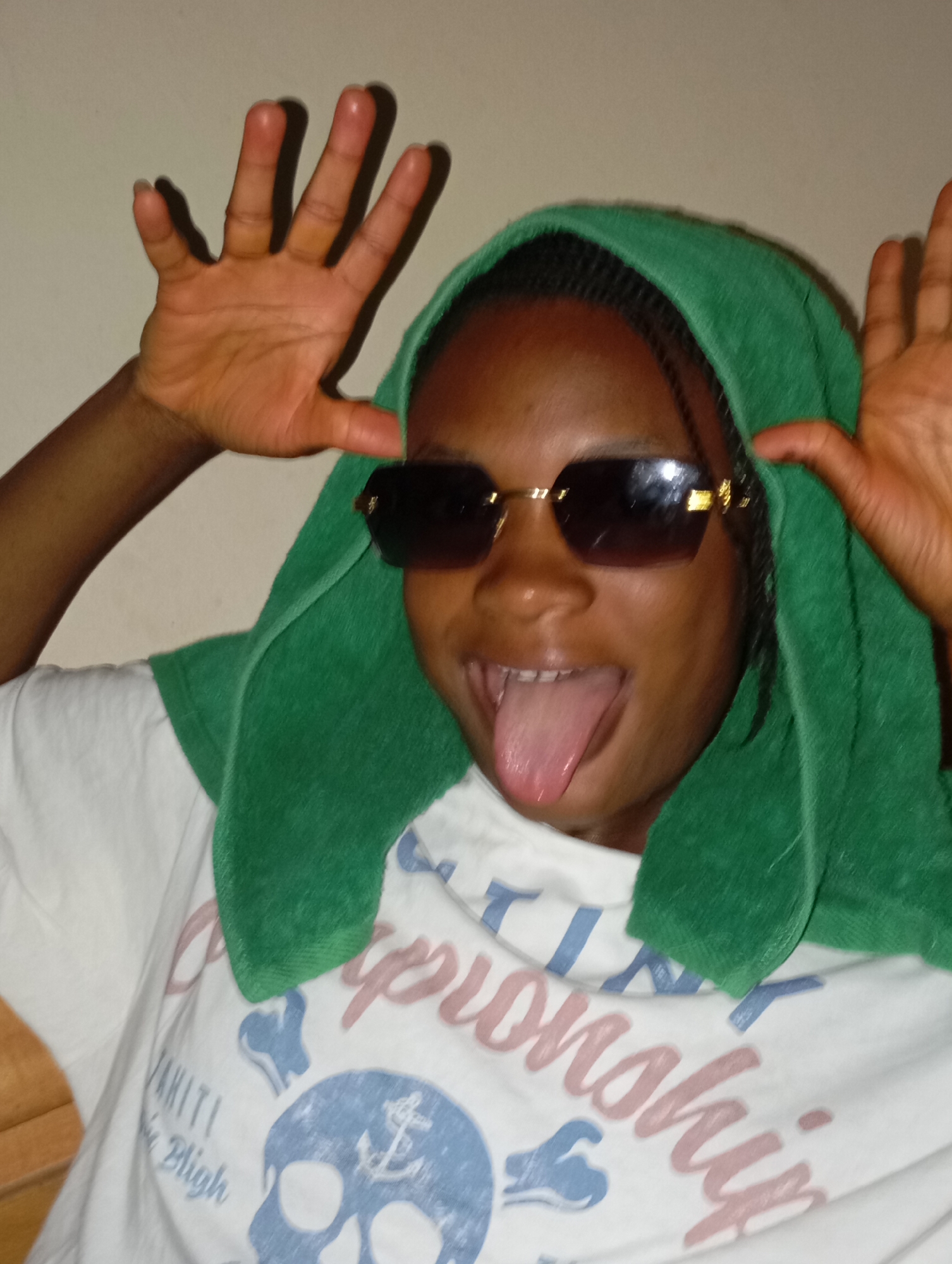A young, dark-skinned individual is posing indoors. The person is wearing sunglasses, a green towel draped over their head, and a white t-shirt with a skull and text design. Their tongue is sticking out, and both hands are raised near their head in a playful manner. The background is a plain light-colored wall. There is no visible information in the image to confirm the location in the city of Jos, Nigeria.