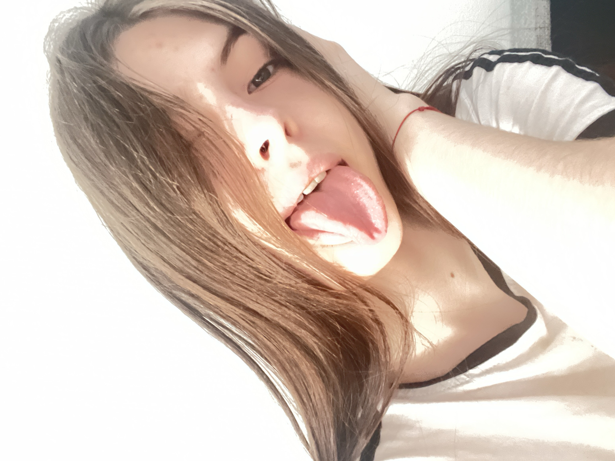 A young female subject is depicted in a close-up, brightly lit indoor setting. Her head is tilted to the right, and she is looking directly at the camera with a playful expression, her tongue extended from her mouth. She has long, straight brown hair with lighter highlights, parted roughly in the middle, with a portion obscuring her left eye. Her right eye is fully visible. She is wearing a light-colored (likely white) t-shirt with a dark-colored V-neck trim. On her right shoulder, three dark stripes are visible, indicative of athletic wear. A thin red string bracelet is present on her right wrist, and a small mole is visible on the right side of her neck near the collarbone. Her right hand is raised, with fingers partially obscured by her hair on the right side of her head. Strong, natural light illuminates the scene from the upper left, casting distinct highlights on her skin and hair and soft shadows, particularly under her chin. The background is a plain, bright white wall, suggesting a clean, minimalist indoor environment. The specific location in the city of Saint Petersburg, Russia, cannot be determined from the visual information presented in the image.