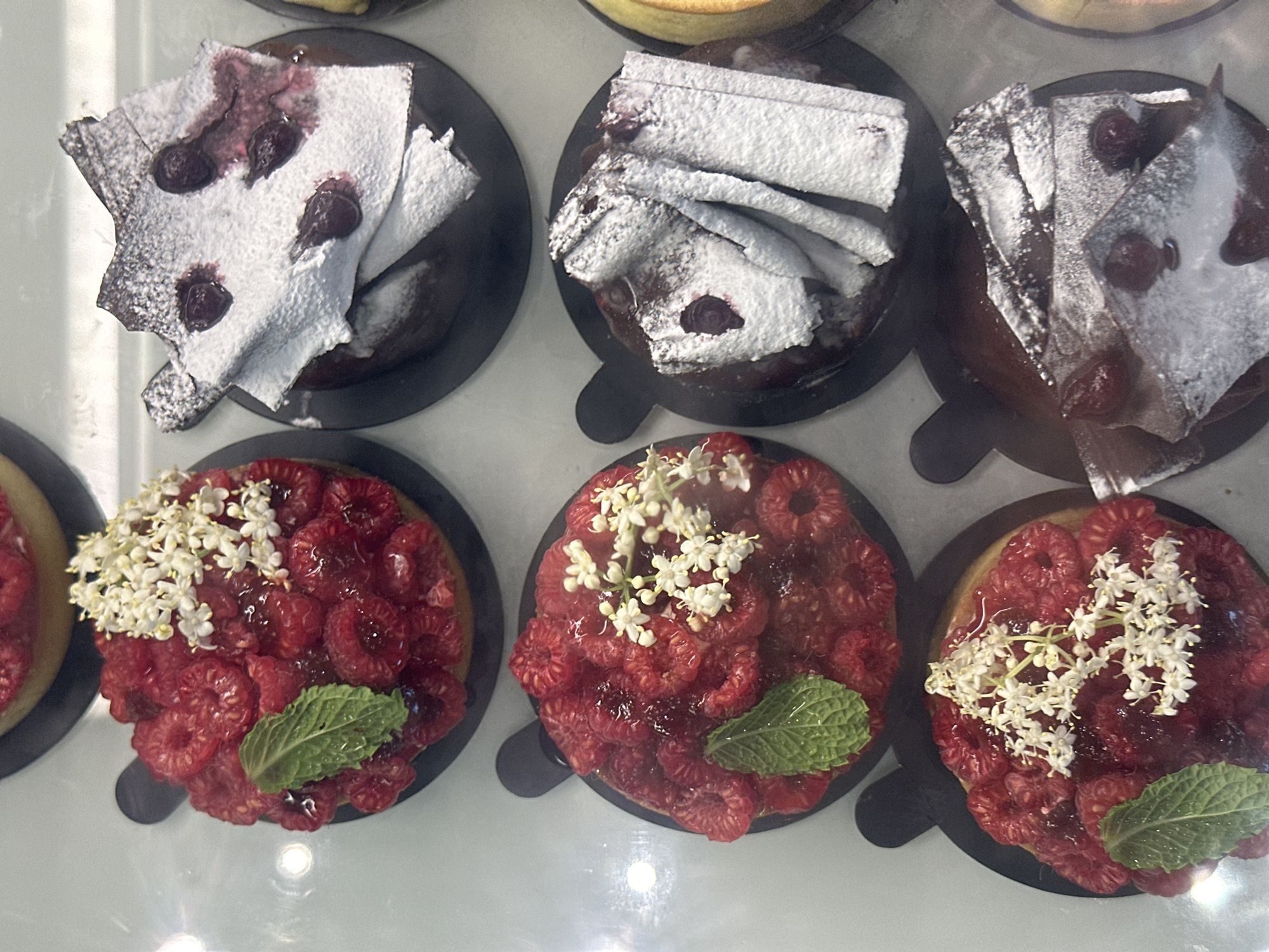 Six pastries are displayed in a refrigerated case. The pastries are arranged in two rows of three. The pastries in the top row are on the left and right, with the center pastry mostly obscured. The pastries in the bottom row are clearly visible. From left to right, the bottom row contains a raspberry tart with a mint leaf and white flowers, a chocolate pastry topped with powdered sugar and cherries, and another chocolate pastry with similar decoration. The top row appears to have a similar raspberry tart on the left and a chocolate pastry on the right. The pastries are on black circular bases. The image is taken from above, looking down into the display case. The background is a blurred white surface. The scene is likely in a bakery or patisserie in North Kuta, Indonesia.