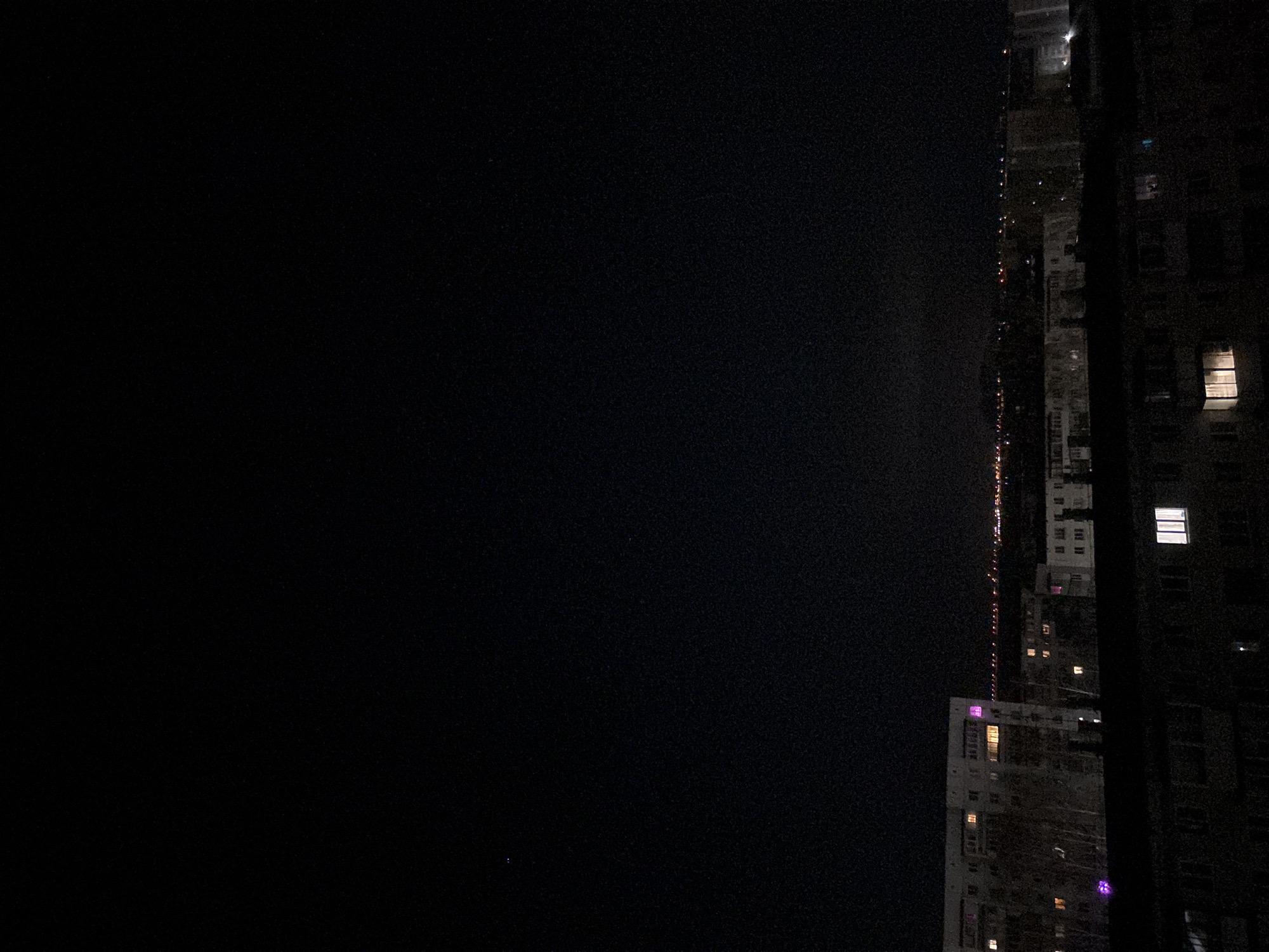 The image depicts a nighttime scene in an urban environment, likely in Chaikovsky, Russia. On the right side of the frame, there are several tall buildings, possibly apartment complexes or office towers. Some of the buildings have illuminated windows, indicating activity inside. A smaller building near the bottom of the frame seems to be lighted with a purple hue. To the left, a dark expanse represents either the night sky or a shadowed area between buildings. Thin lines of light, probably streetlights, are visible along the lower-center area of the frame, suggesting a distant road or street. The overall impression is that of an urban night scene, captured with a focus on the contrasting light and dark elements of the city's landscape.