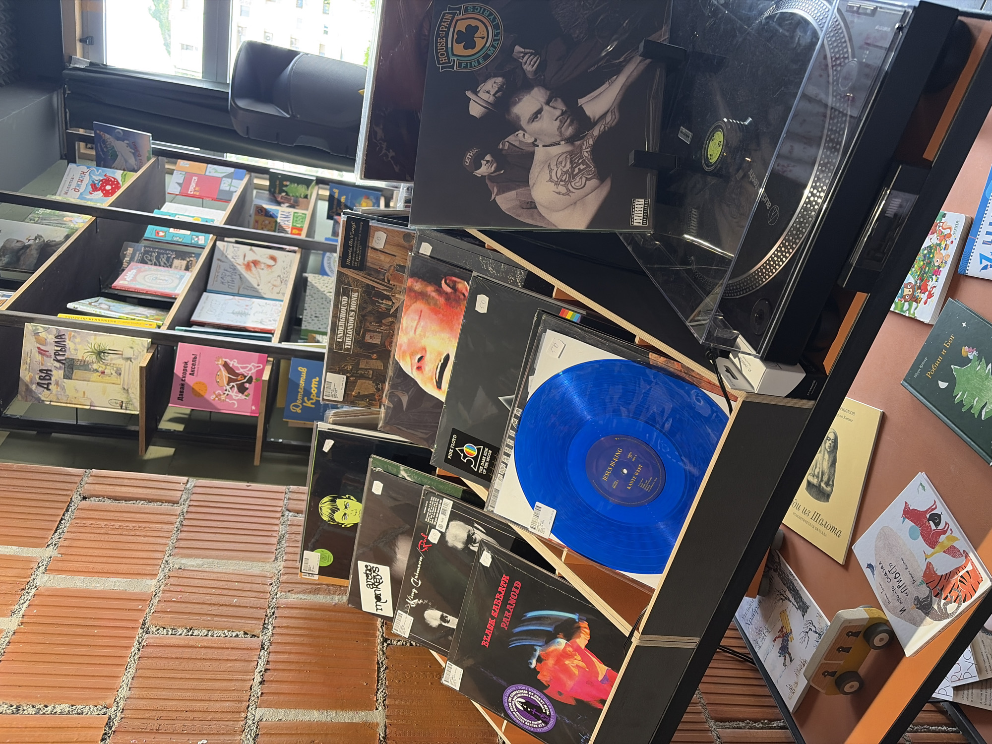 This is an indoor shot, likely in a retail space in Budva, Montenegro, featuring a display of vinyl records and children's books. The display shelves are made of black metal and wood, arranged diagonally against a brick wall. A turntable sits atop the vinyl display. Vinyl records from bands like House of Pain, Pink Floyd, King Crimson, and Black Sabbath are visible. Children's books are on shelves to the left and right. The lighting appears to be a mix of natural light from a window visible in the background and possibly indoor lighting. The floor is brick.