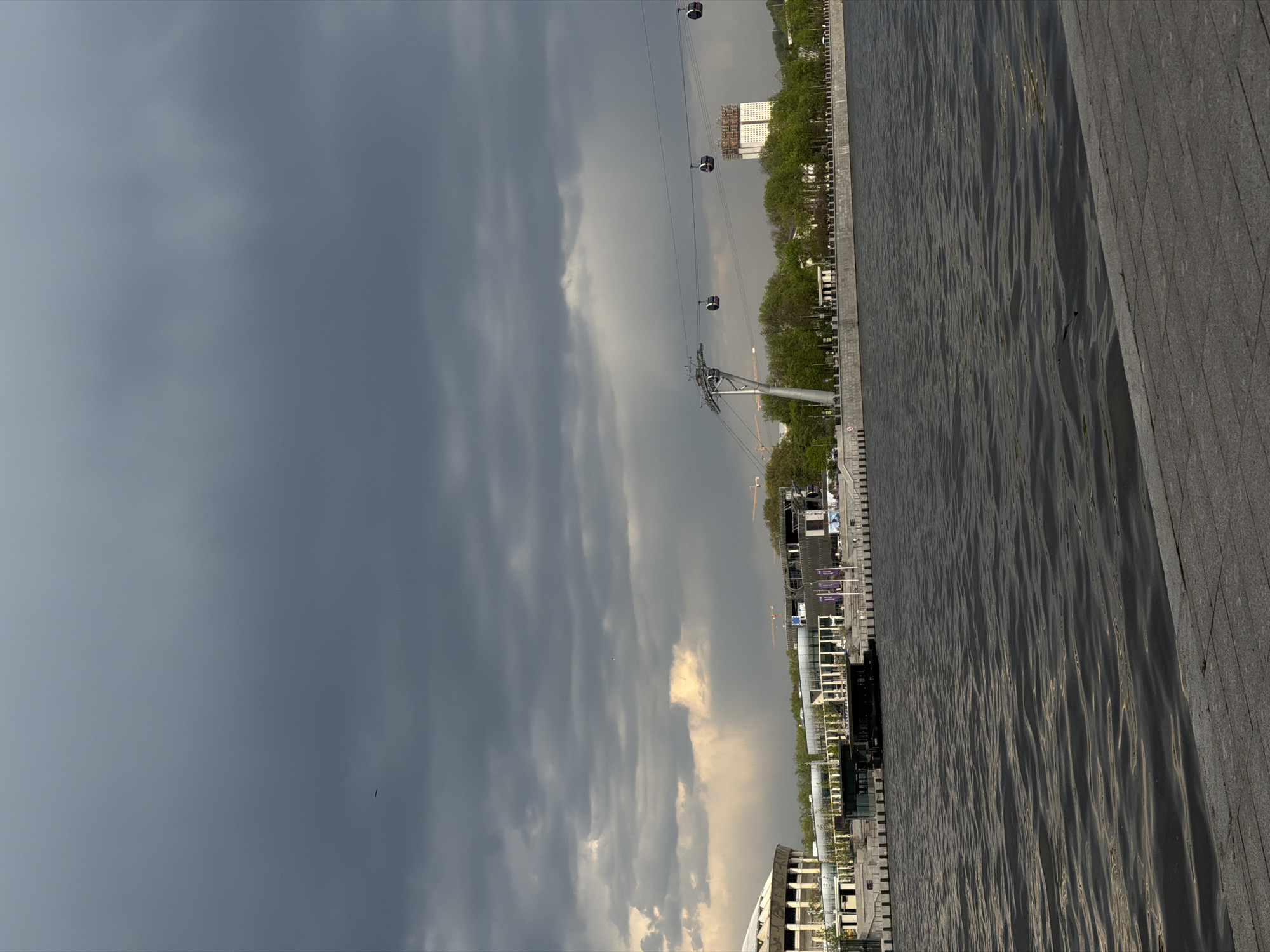 The image depicts a landscape in Moscow, Russia, showing a section of the embankment and the Moskva River under a cloudy sky. A cable car system stretches above the embankment with support towers visible, and a row of trees lines the riverbank. Modern buildings and architectural structures are along the embankment. The foreground features a paved area. The sky is overcast with dark clouds.
