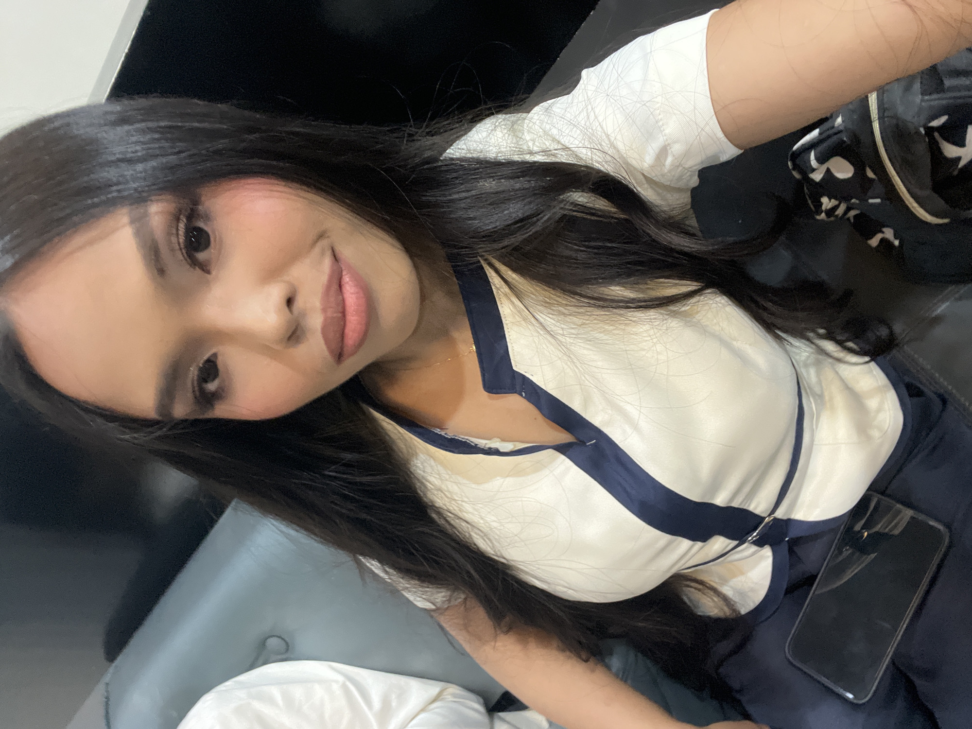 The image shows a selfie of a woman with long, dark hair, wearing a white and blue collared shirt. She is seated on a dark-colored couch, possibly in a waiting area or lounge. A smartphone is visible on her lap, and a black and white patterned bag is next to her. The background is dark, suggesting an indoor setting. The woman appears to be in a relaxed pose, looking towards the camera. Based on the context of the image being provided, the location is identified as Davao City, Philippines.