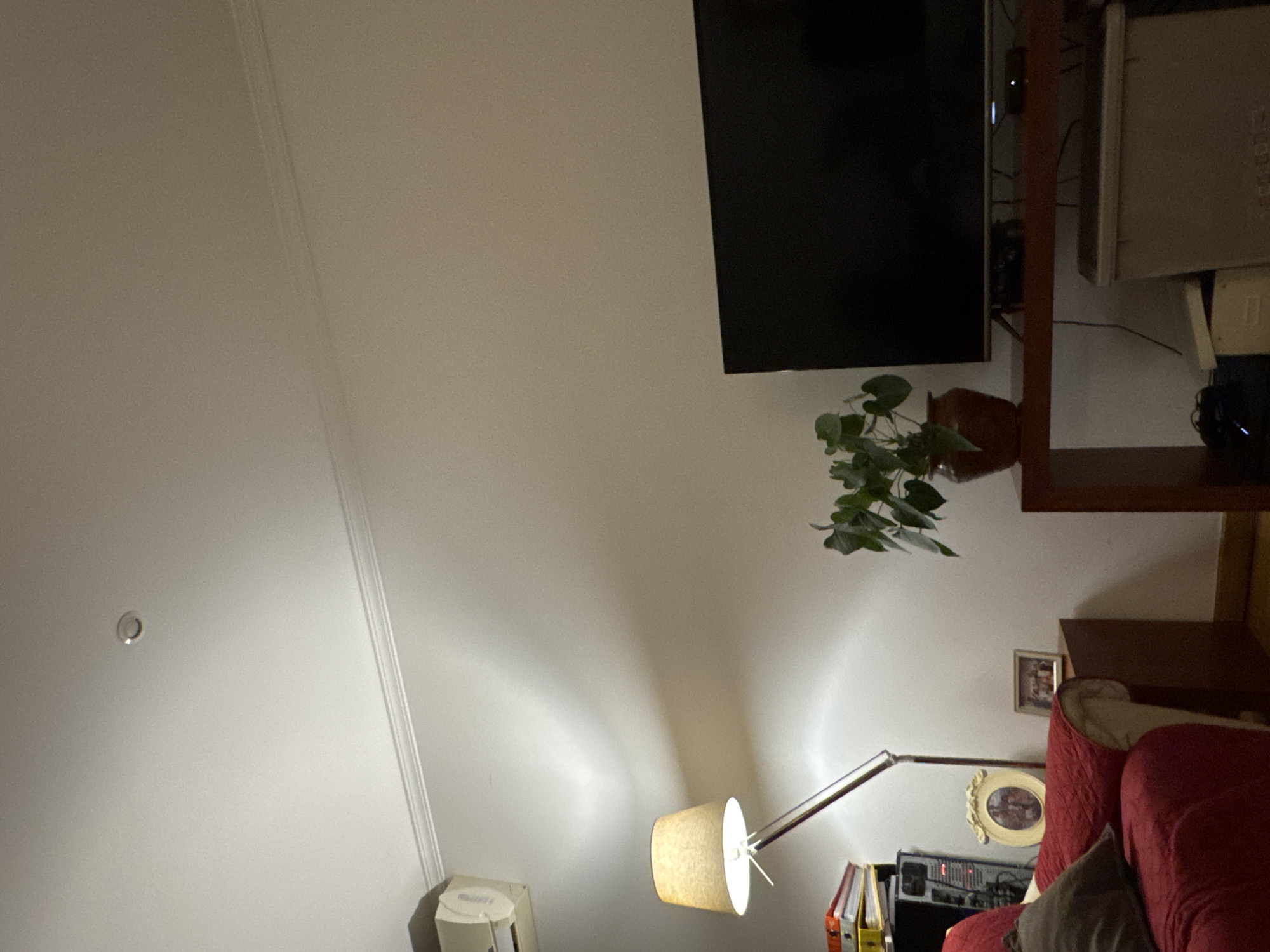 The image depicts an interior residential living space. The specific location within Mirandela, Portugal, cannot be determined from the visual information provided. The scene is illuminated by artificial light sources, including a floor lamp with a beige fabric shade on the left and a single recessed light fixture in the ceiling. The walls are light-colored with visible crown molding, and a white air conditioning unit is mounted high on the left wall.
In the lower-left quadrant, a red armchair or sofa is partially visible, covered with a quilted red throw and a dark cushion. Adjacent to it, a wooden side table holds an oval, ornate framed photograph and a smaller, rectangular framed photograph. Behind the floor lamp, a stack of binders or files is present next to a stereo system component.
A dark brown console table dominates the center-right, supporting a large, black, flat-screen television with an unlit screen. A potted plant with green leaves in a brown pot is situated on this table to the left of the television. Beneath the console table, a large, light brown storage bin and other items are present. A wooden floor is visible in the lower right portion of the frame. No individuals are depicted.