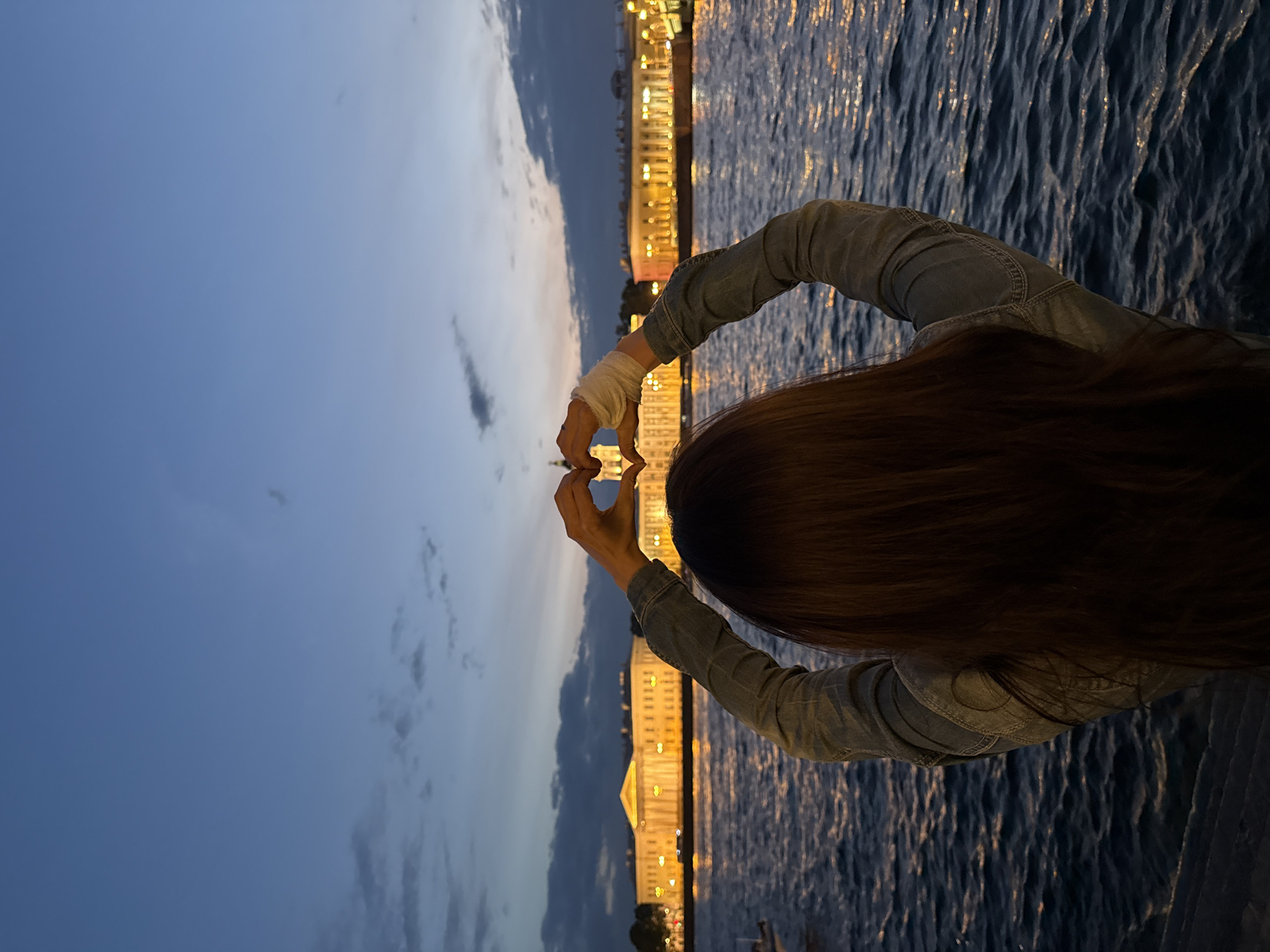 The image, captured at dusk or twilight, presents a rear view of a person standing by a body of water, likely a river or canal in Saint Petersburg, Russia. The individual has long, dark hair and is wearing a denim jacket. Their arms are raised, and their hands are interlocked to form a heart shape, seemingly framing the distant urban landscape. A white bandage or wrapping is visible on the person's left wrist.
Across the water, a long line of stately buildings with classical architecture is brightly illuminated, their lights casting shimmering reflections on the dark, rippled surface of the water. The sky overhead is a deep blue, indicative of the evening hours, with some soft cloud formations visible. The overall scene conveys a sense of admiration or affection for the illuminated city view. No text is visible in the image.