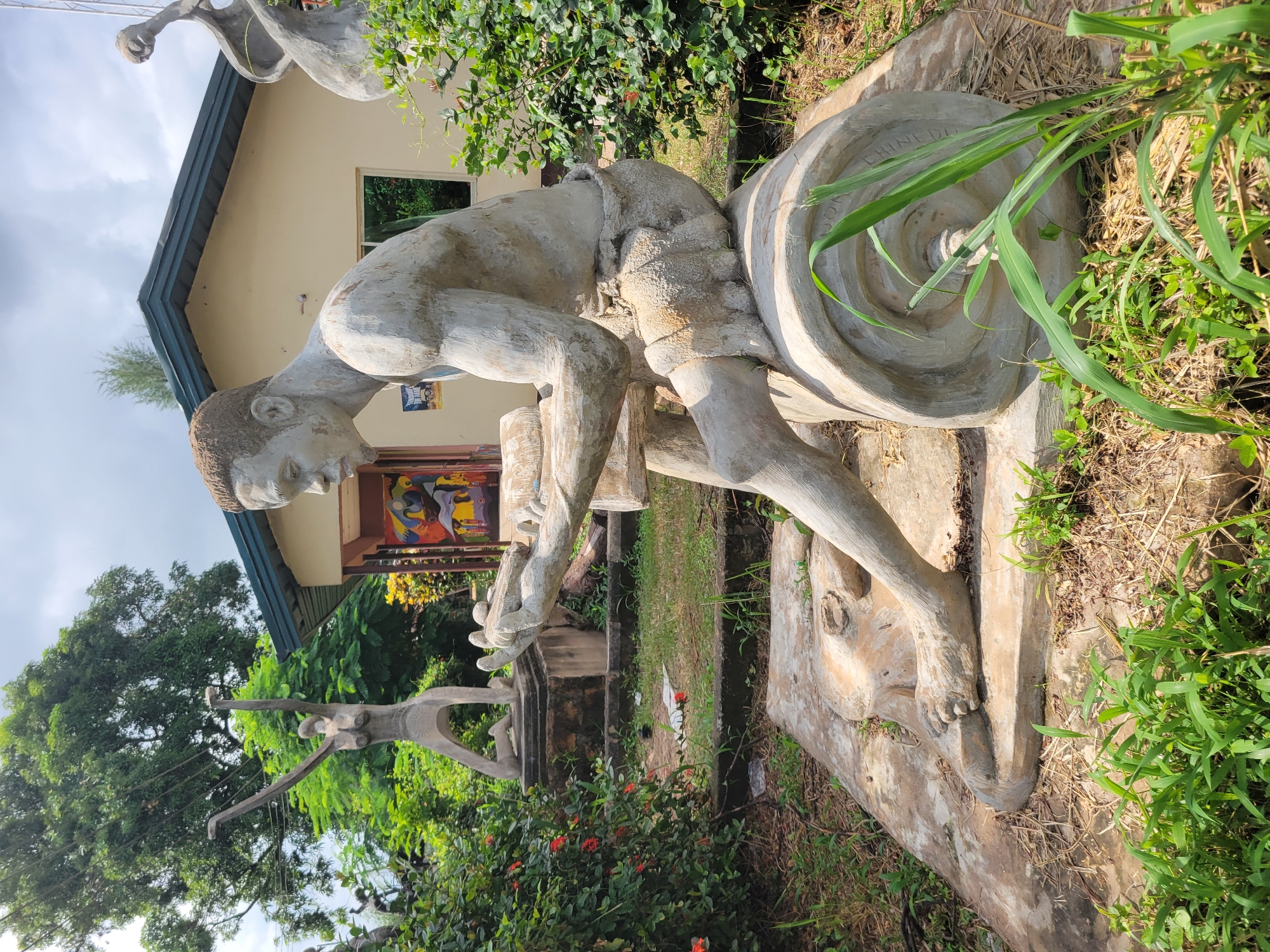 In the city of Egbagu, Nigeria, a concrete statue of a man is seated on a large concrete cylinder. The statue depicts the man with short, textured hair, an athletic build, and wearing a cloth around his waist. He is holding a cylindrical object in his hands, appearing to be engaged in a task. The statue is located outdoors, in a grassy area with bushes and trees surrounding it. In the background, a building with a light-colored facade and a dark roof is visible. To the left of the main statue, another concrete statue of a female figure in a dynamic pose with arms raised is present. The scene suggests an outdoor art installation or sculpture garden.