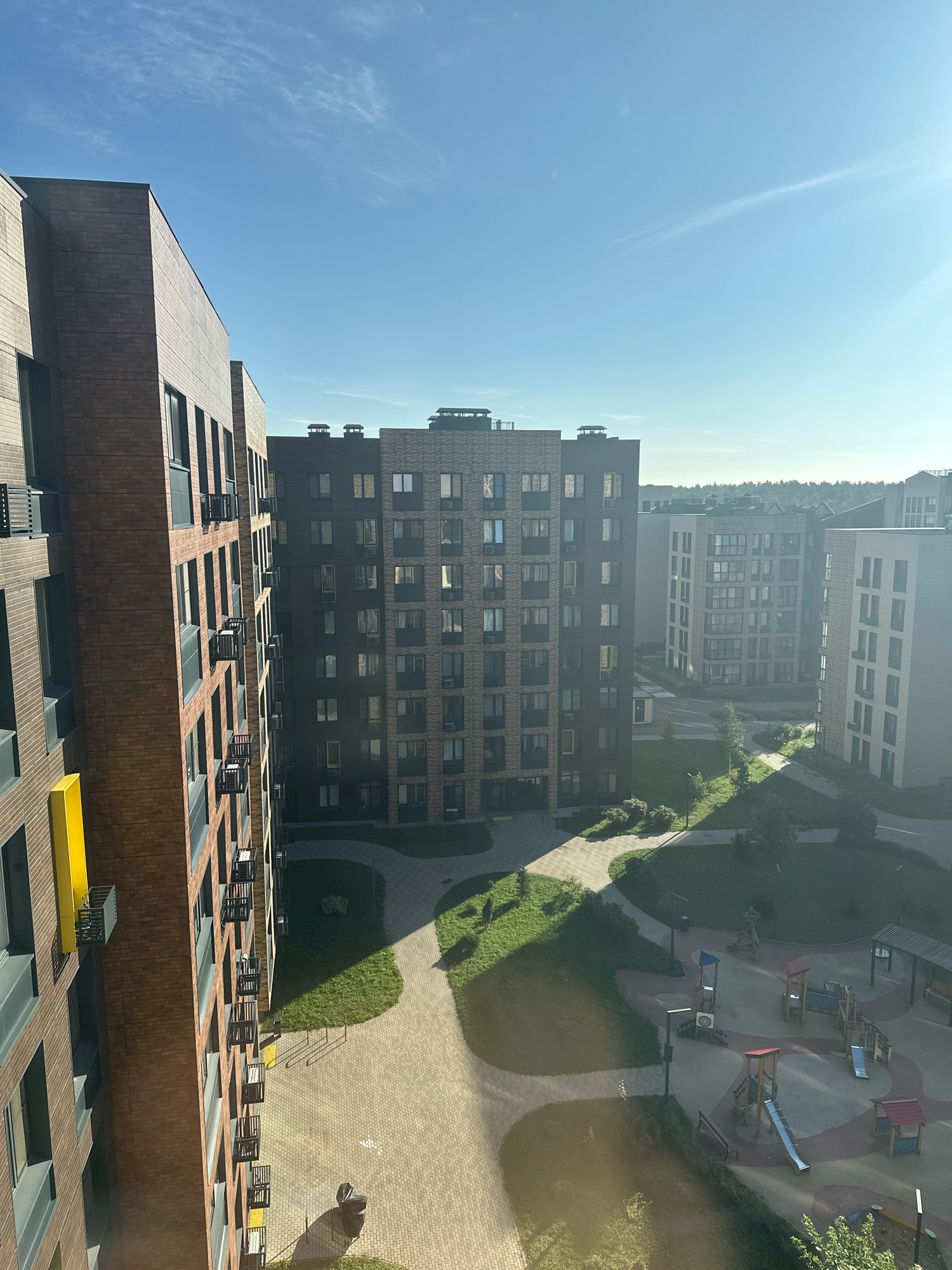 An aerial view captures a modern residential complex in Vidnoye, Russia, under a clear blue sky with faint wispy clouds. The foreground features a tall, multi-story building constructed with dark brown brick, featuring numerous windows and small black balconies. To its right, a slightly shorter building of similar design with lighter brown brick is visible, also with many windows and balconies. Further in the distance, a row of similar residential buildings stretches across the landscape. The ground level of the complex includes a playground with various colorful structures like slides and climbing frames, surrounded by paved pathways and patches of green grass and trees. The scene is bathed in sunlight, casting long shadows across the courtyard and highlighting the architectural details of the buildings and the playground.