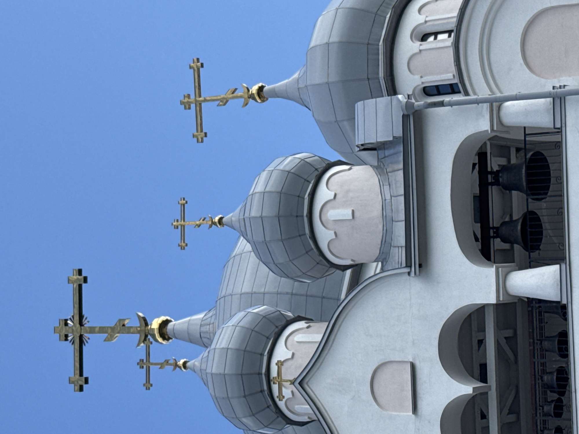 The image shows the upper part of a white Orthodox church with multiple domes and crosses against a clear blue sky. The domes are silver-colored and have a segmented design. Several golden crosses are visible, some with a distinct Orthodox design featuring an additional bar and crescent moon at the bottom. Arched openings revealing large bells are visible on a lower level of the church structure. The architectural style suggests a historical religious building. The specific location in Saint Petersburg is not identifiable from the image alone. No people are present in the scene.