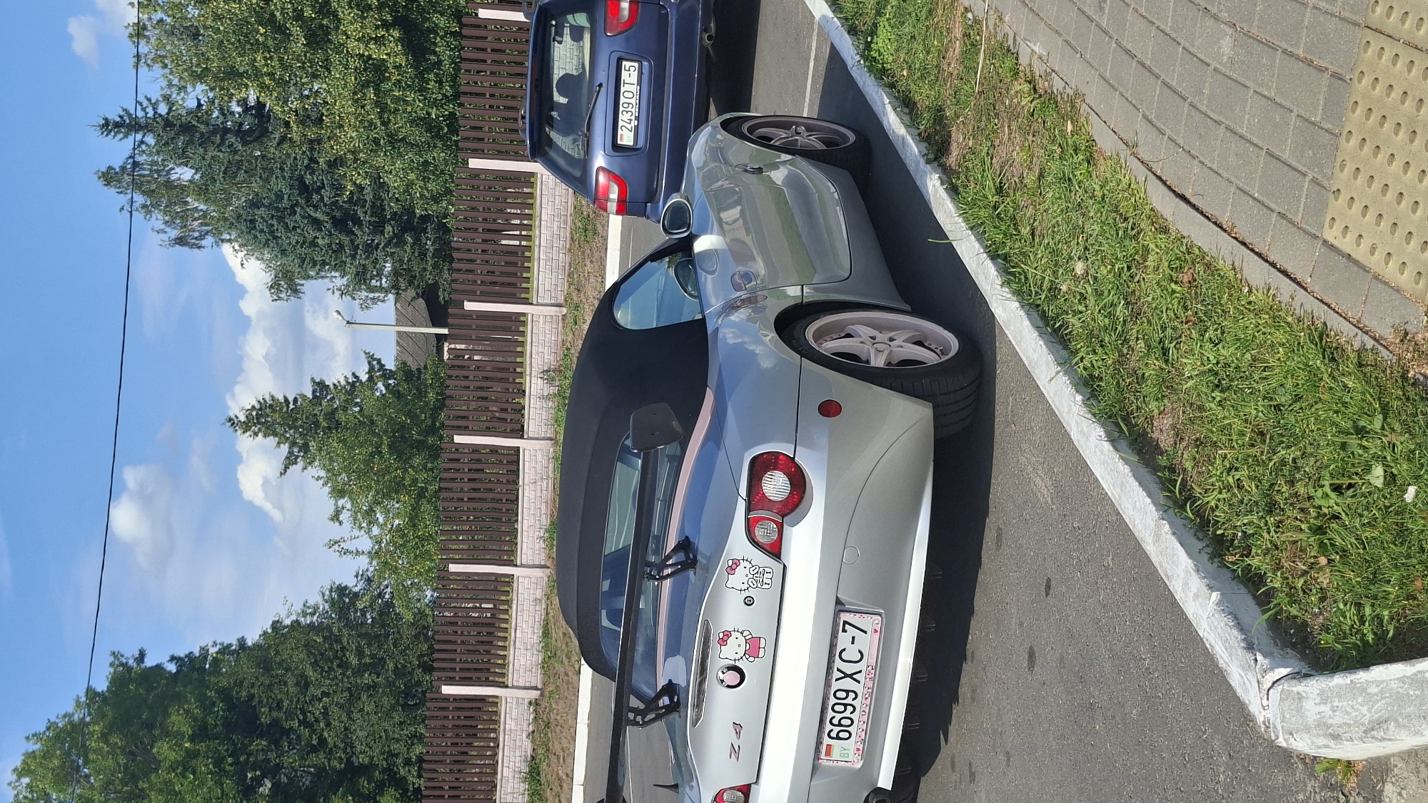 A silver BMW Z4 convertible with a black soft top is parked on an asphalt surface next to a blue Audi A4 wagon. The BMW has a large black rear spoiler and is decorated with two Hello Kitty stickers on the trunk. Its license plate reads "6699 XC-7 BY". The Audi's license plate reads "2439 OT-5". Both vehicles are parked in what appears to be a residential or commercial area with a wooden fence and trees in the background. The scene suggests a warm, sunny day. The location is identified as Myadzyel, Belarus.