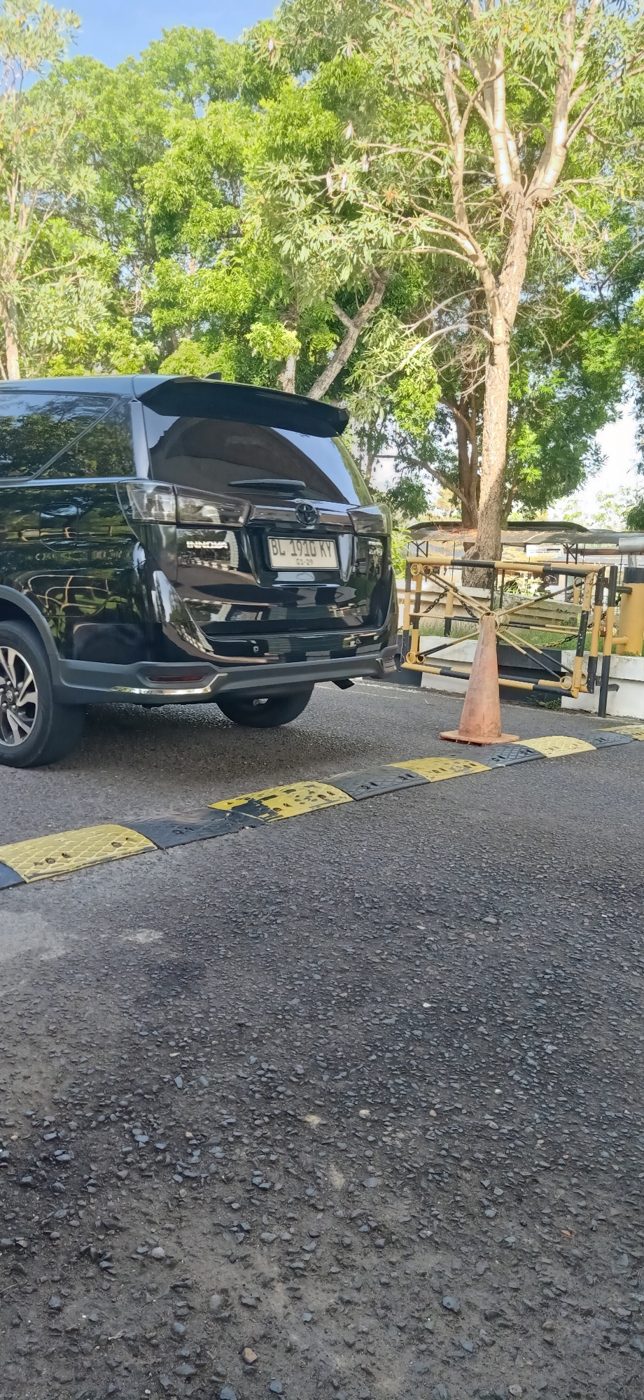 A black Toyota Innova is parked on an asphalt road in Tambontunong, Indonesia. The vehicle's rear is facing the camera, and its license plate reads "BL 1910 KY". The car is positioned behind a speed bump made of alternating black and yellow sections. To the right of the car, there's a yellow and black striped barrier gate and an orange traffic cone. In the background, lush green trees with visible branches and leaves create a natural backdrop under a clear blue sky. The overall scene depicts an outdoor parking area with a focus on the vehicle and its immediate surroundings.