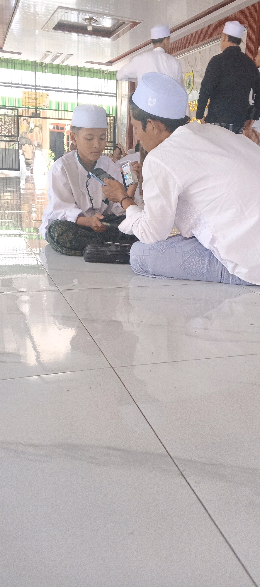 Two young males, appearing to be Indonesian, are seated on a tiled floor indoors. The male on the left is wearing a white long-sleeved shirt, a green and white patterned sarong, and a light blue peci (cap). He is looking down at a smartphone in his hands. The male on the right is wearing a white long-sleeved shirt and light blue patterned pants, also with a light blue peci. He is also looking at a smartphone and holding a small white packet. In the background, other individuals are visible, also wearing traditional Islamic attire including peci. The environment appears to be a communal gathering space, possibly a mosque or a hall, with reflective tiled flooring and a white ceiling. The overall lighting suggests it is daytime. The image's context is not explicitly stated to be Sungelebak, Indonesia.