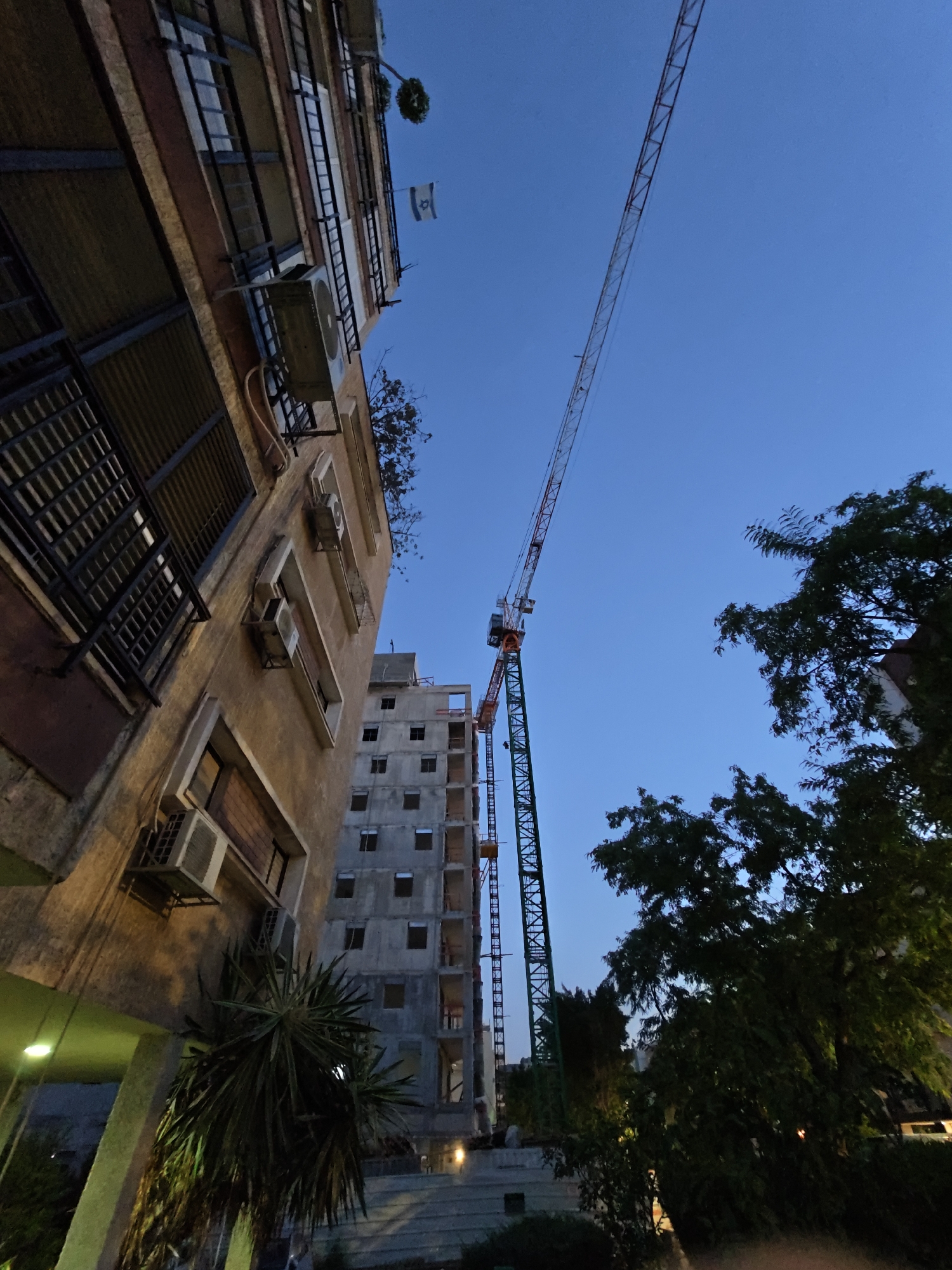 The image captures a construction site at dusk in Petah Tikva, Israel. The foreground features a building with balconies and air conditioning units, while a tall construction crane dominates the sky. Adjacent to the existing building, a new structure is under construction, showing concrete framing and window openings. Trees and foliage are visible in the mid-ground and right side of the image. An Israeli flag is visible on the balcony of the existing building. The overall scene depicts urban development in progress under a clear blue sky.